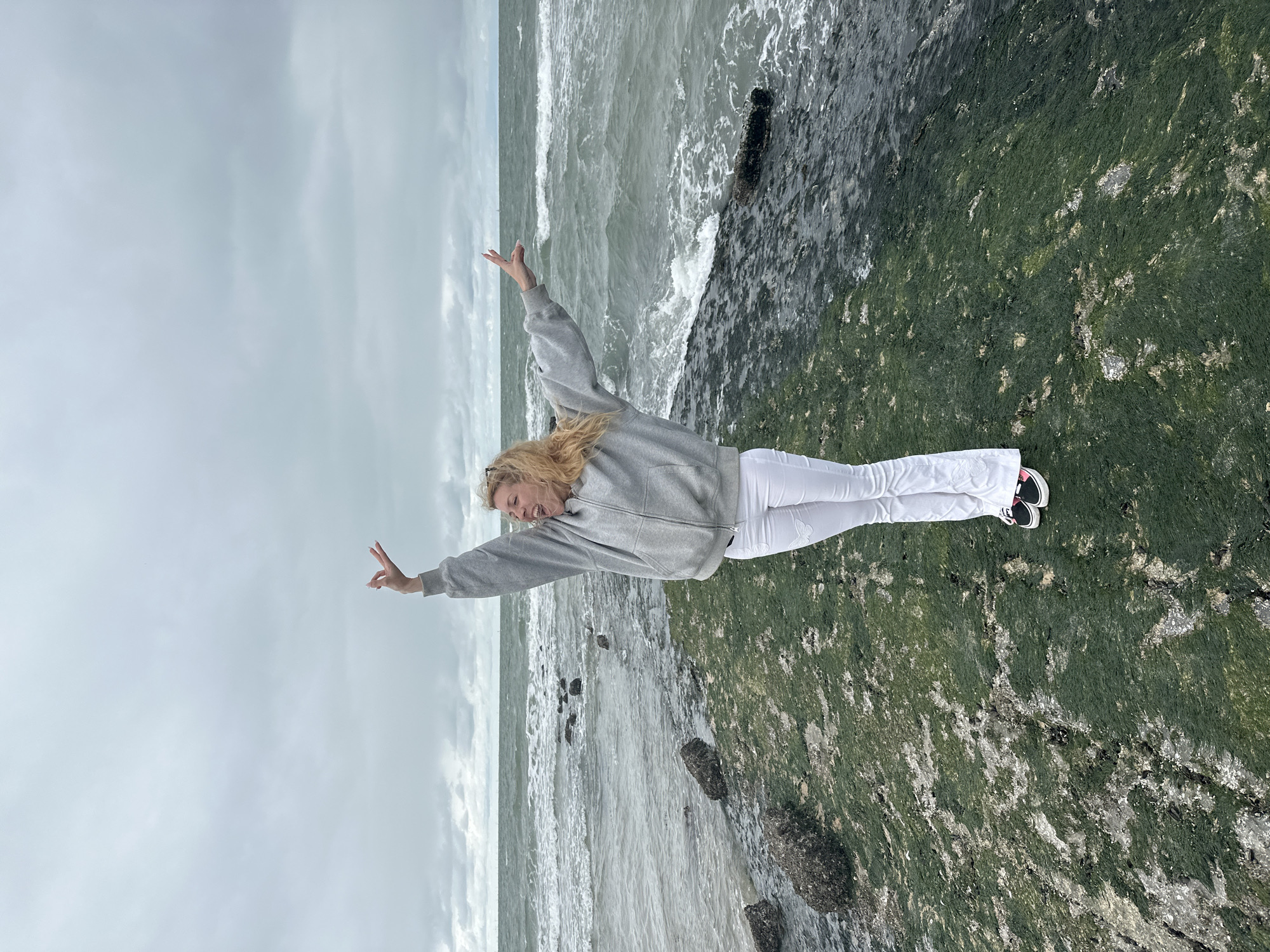 A woman with blonde hair stands on a rocky, algae-covered shore, facing the sea. She is wearing a grey hooded sweatshirt and white flared pants. Her arms are raised in a gesture of joy or excitement, and her mouth is open as if she is shouting or laughing. The sea is choppy with waves, and the sky is overcast. The location is Knokke-Heist, Belgium, a coastal resort town.
