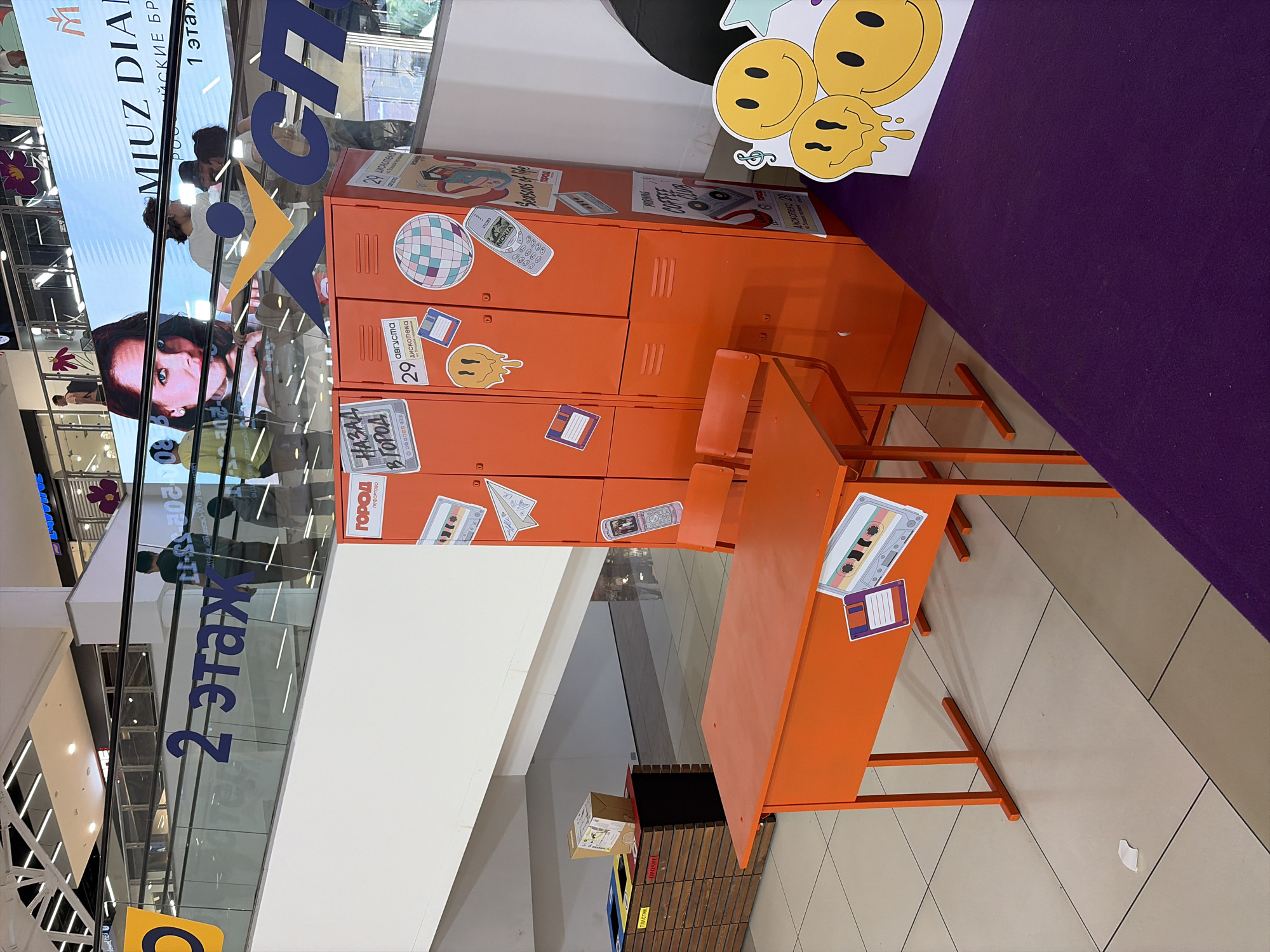 The image depicts an interior scene within a modern shopping mall, likely in Moscow, Russia, based on the Cyrillic text visible on signs and displays. In the foreground, an orange school desk with two chairs is set up, adorned with retro-themed stickers such as cassette tapes and floppy disks. Behind the desk, a row of bright orange lockers stands, also decorated with similar vintage-inspired decals. To the left of the lockers, a wooden multi-compartment waste sorting bin is visible, labeled with Cyrillic text indicating "PLASTIK" and "PROCHEY" (likely referring to different types of recyclable materials). In the background, an escalator runs upwards, with several individuals on it, and a large digital billboard displays an advertisement featuring a woman's face and Cyrillic text, including "MIUZ DIAMOND" and pricing information. The overall environment suggests a themed display or event within the mall, possibly related to fashion, entertainment, or a nostalgic theme.