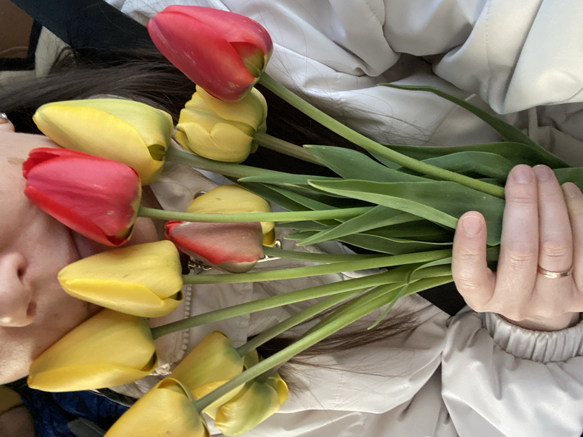 A person, possibly female, lies on their back, holding a bouquet of tulips. The tulips are a mix of red and yellow. The person's hand holding the bouquet has a gold ring on the ring finger. The background includes a light-colored jacket or coat. No information indicates the location is Chaikovsky, Russia.