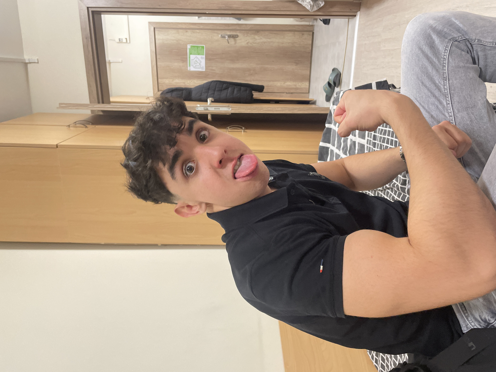 A young man with curly dark hair is posing for a photo while sticking his tongue out. He is wearing a dark polo shirt and light gray jeans. He is sitting on a bed with a black and white patterned sheet. The room has light brown wooden furniture, including a wardrobe, desk, and a door leading to another room. There is a jacket and sandals on the desk. The location appears to be indoors, possibly a dormitory room. The city of Sofia, Bulgaria, cannot be determined from the image alone.