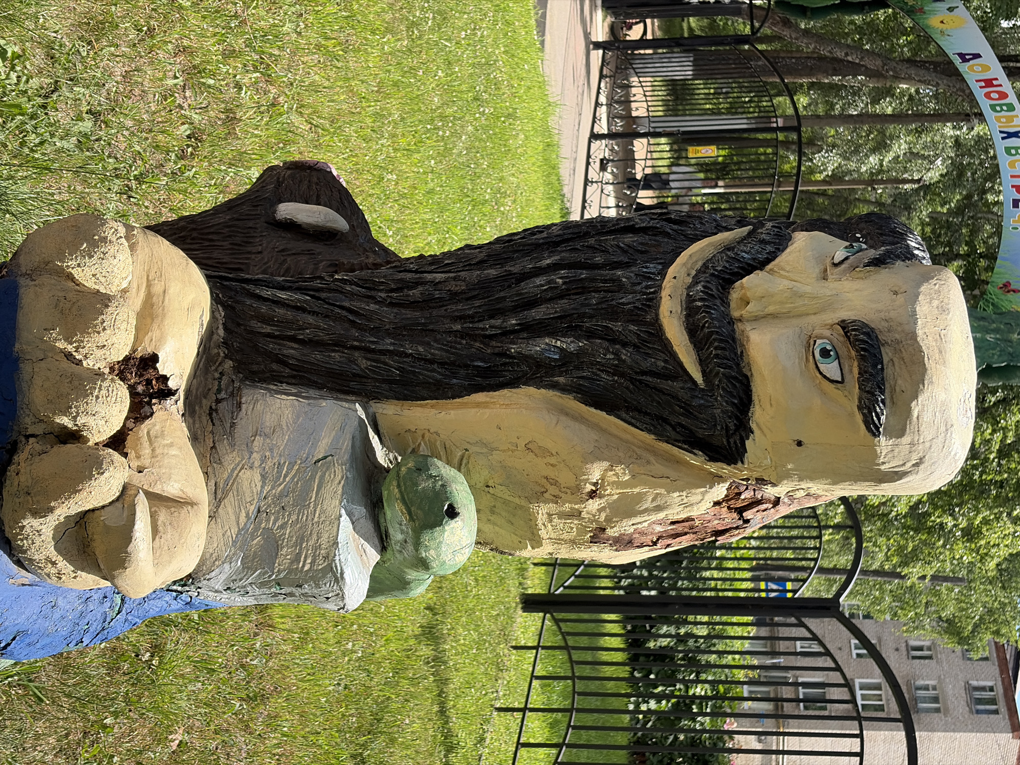 This media file depicts a close-up, rotated view of a wooden sculpture of a man, likely a fictional character, with a large, dark beard and mustache. The sculpture is painted in various colors, including beige for the face and hands, dark brown for the hair, and a light blue for the base. The man's eyes are painted a striking blue. He appears to be holding a green object, possibly a turtle, in his left hand.
The sculpture is situated outdoors, with a background of green grass, a black metal fence, trees, and buildings. The lighting suggests it is daytime, likely sunny, given the shadows. A curved banner with text in Russian is visible in the upper right quadrant of the image.
The text on the banner reads "ДО НОВЫХ ВСТРЕЧ" which translates to "Until new meetings." The overall impression is of a whimsical, outdoor art installation, perhaps in a park or public space in Chaikovsky, Russia, designed to be a playful attraction. The sculpture shows signs of age and wear, with chipping paint and exposed wood, adding to its character.