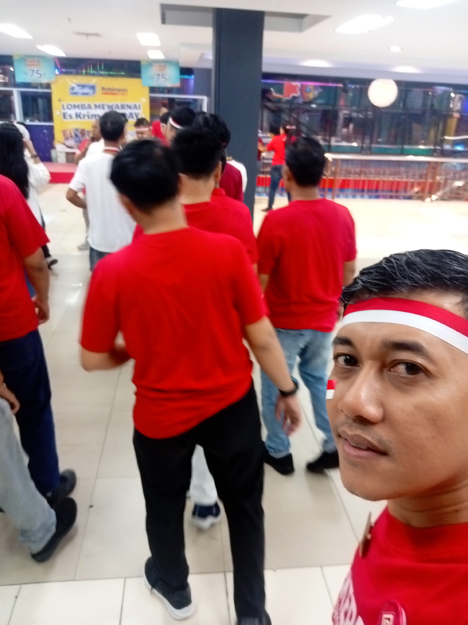 The image shows a group of people in red shirts inside a well-lit indoor space, possibly a mall or an event venue in Batam, Indonesia. A banner in the background reads "LOMBA MEWARNAI Es Krim RAY," indicating a coloring competition for ice cream. The man in the foreground is wearing a red and white headband, possibly a participant or organizer. The overall atmosphere appears to be festive and engaged.