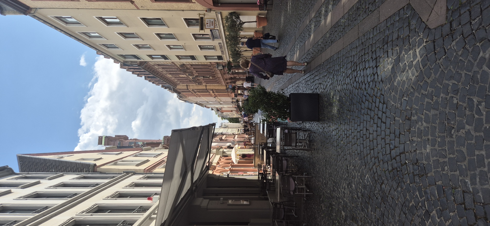 This image captures a street scene in Mainz, Germany, likely in the historic city center, given the cobblestone paving and the architecture. The perspective looks down a narrow street lined with multi-story buildings that exhibit a mix of traditional European styles with light-colored facades and ornate detailing on some.
In the foreground on the left, an outdoor seating area of a cafe or restaurant is set up with dark metal tables and chairs arranged on the cobblestones. A dark awning extends over this area. To the right of the cafe seating, a large dark planter box contains a dense green shrub.
Further down the street, other tables and chairs are visible, suggesting a bustling area with dining establishments. Several people are walking along the street. Notably, a woman in a dark patterned dress with a beige shoulder bag is walking away from the camera towards the background on the right side of the street. Behind her, a man in a dark shirt and blue jeans, carrying a dark bag, is also walking away from the camera. More people are scattered further down the street, indicating activity and pedestrian traffic.
The buildings on both sides of the street have numerous windows and decorative architectural elements. The sky above is partly cloudy with patches of blue sky. The cobblestone street is a prominent feature, showing a pattern of dark, interlocking stones. The overall atmosphere suggests a pleasant day in a European city.