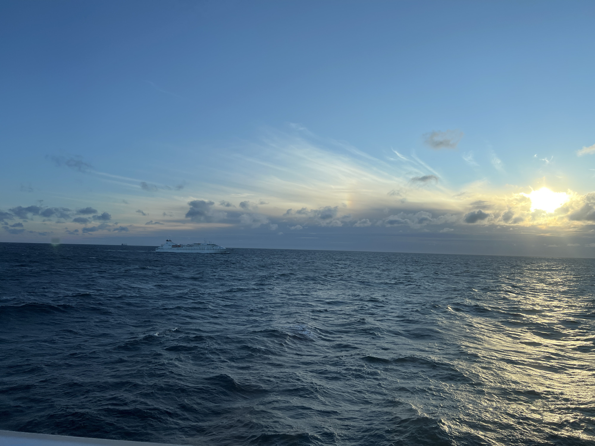 This is a horizontal, eye-level, outdoor, full shot of a large, white cruise ship sailing on a deep blue sea under a partly cloudy sky. The sun is setting or rising on the right side of the image, casting a warm, golden light on the water and clouds. The sea is choppy with moderate waves, and the sky is a gradient of blue, with wispy clouds and darker clouds lower on the horizon. The cruise ship is in the distance, on the left side of the frame, and appears to be moving from left to right. There are no people visible in the image. The location is unknown, but the image depicts a serene maritime scene.