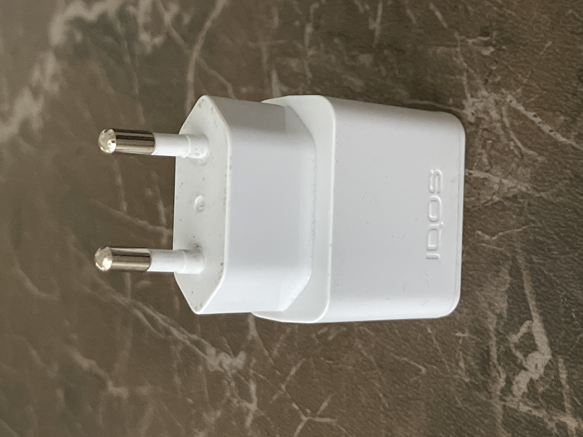 A white IQOS wall charger is placed on a brown, marbled surface. The charger has two metallic prongs extending from the top. The brand name "IQOS" is embossed on the front of the lower section of the charger.
