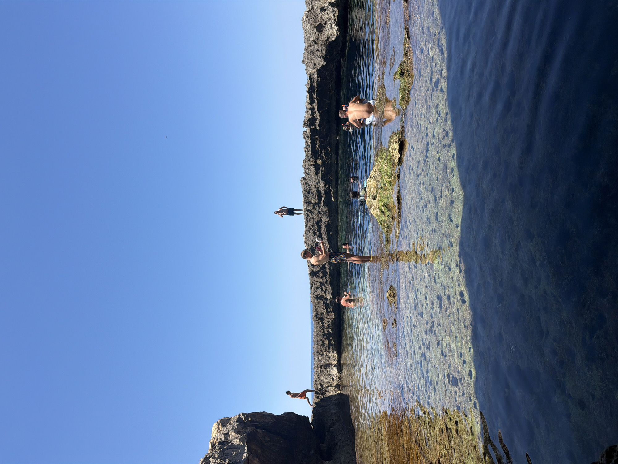 The image depicts a natural rocky swimming pool on a sunny day in the locality of Saint Lawrence, Malta. Several individuals are present in and around the water. In the foreground, the clear, shallow water reveals the rocky seabed. To the left, a large rock formation juts out over the water, with a person in orange shorts walking along its edge. Further back, a man in the water appears to be holding something. Several other people are visible swimming or standing in the pool, and a person stands on a rocky outcrop in the background. The sky is a clear, bright blue, and the sea beyond the pool is also visible.