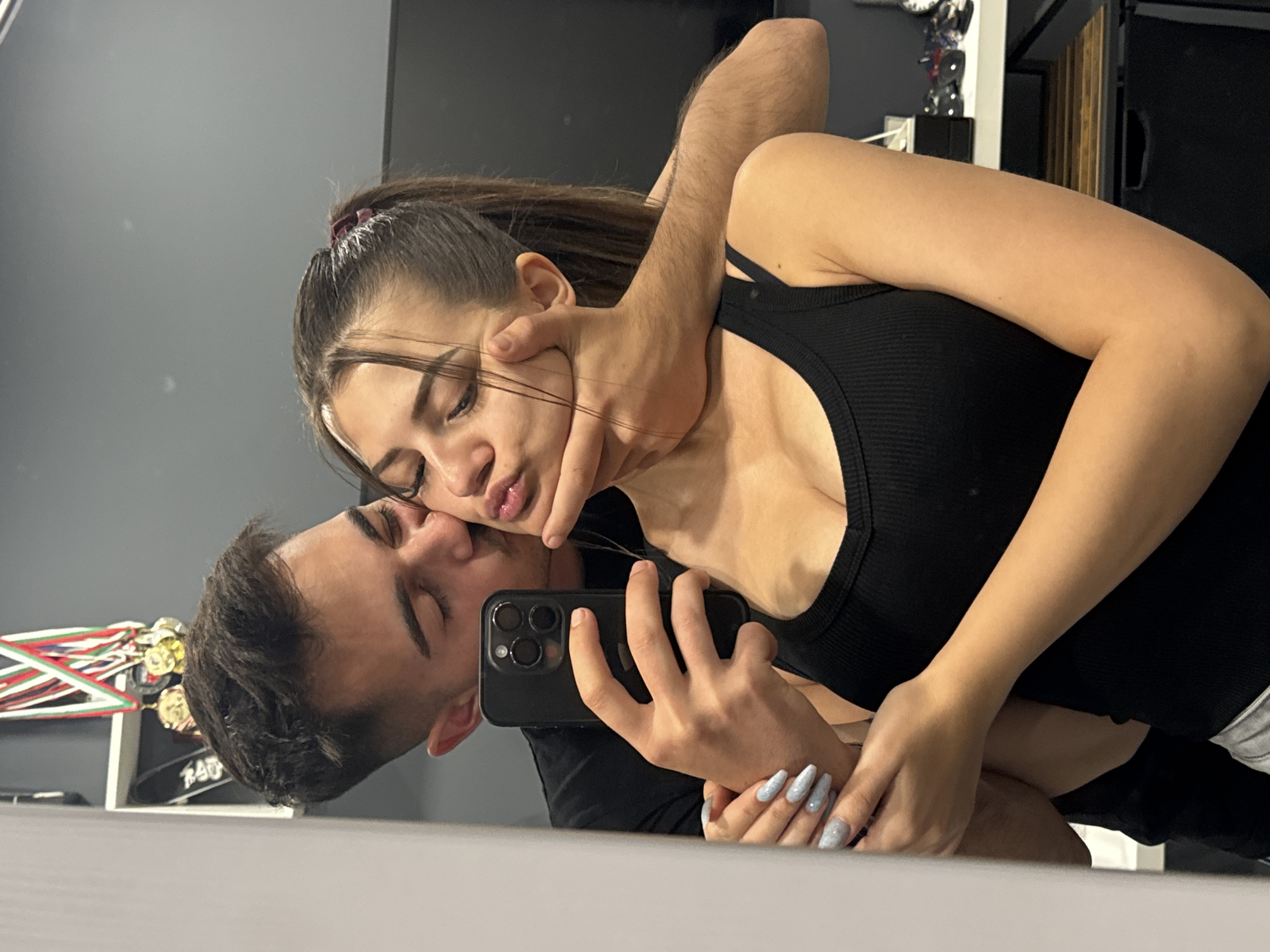 The image shows a man and a woman in an embrace, reflected in a mirror. The woman, with her hair pulled back and wearing a black tank top, has a man's arm around her neck. The man is taking a selfie with a black smartphone, his face close to hers, and his eyes closed. His other arm is around her waist, and her hands are resting on his arm. The background reflection shows part of a shelf with decorations and a closed door. The location in Sofia, Bulgaria, cannot be determined from the image.
