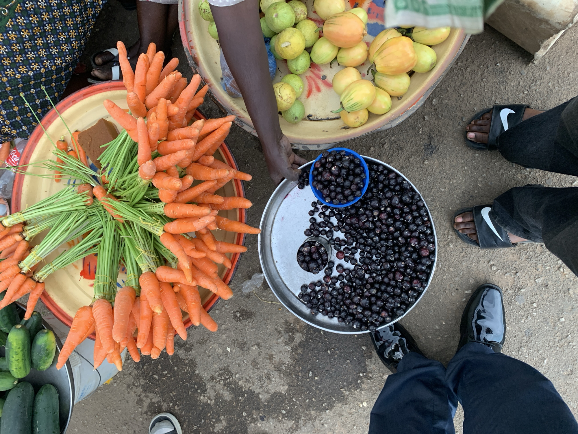 At a street market near New Stadium, Nigeria, vendors offer a variety of fresh produce. Bunches of carrots with green tops are bundled together on a red-rimmed tray. Next to them, a round tray holds dark-colored berries, some in a blue cup. A third tray contains yellow and green fruits. Three people's feet are visible. One wears black Nike sandals, another wears shiny black dress shoes, and another's shoe is partially visible. The ground is gray and rough. One vendor's arm is seen reaching toward the berries.
