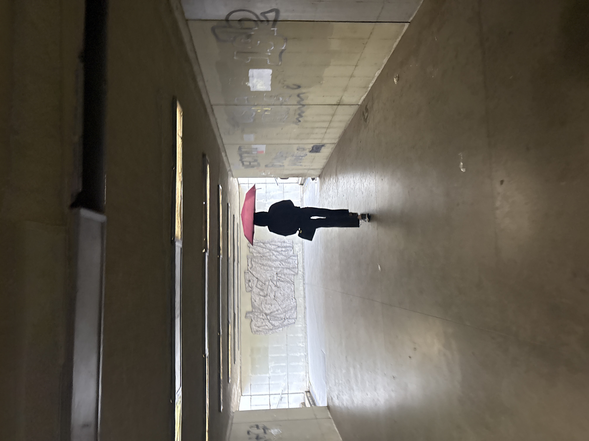 A person walks down a long, narrow concrete tunnel. The person is holding a red umbrella. Graffiti is visible on the walls of the tunnel. The location is most likely in the city of Plovdiv, Bulgaria.