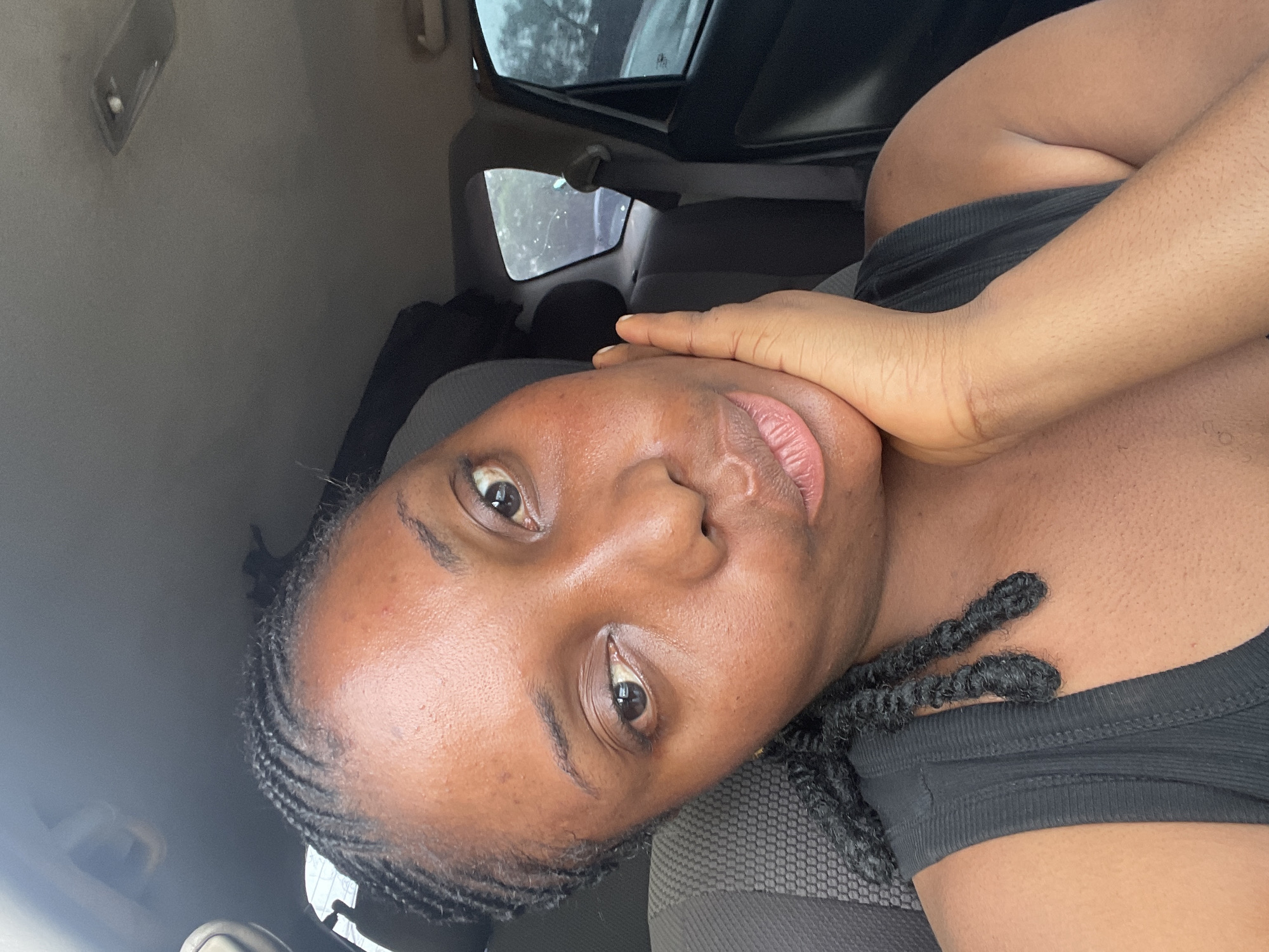 A woman is seated inside a car. She has dark skin and is wearing braids. Her right hand is touching her face. The interior of the car is visible, including the dashboard, seats, and window. The location cannot be ascertained from the image. The city of Jos, Nigeria, cannot be confirmed.