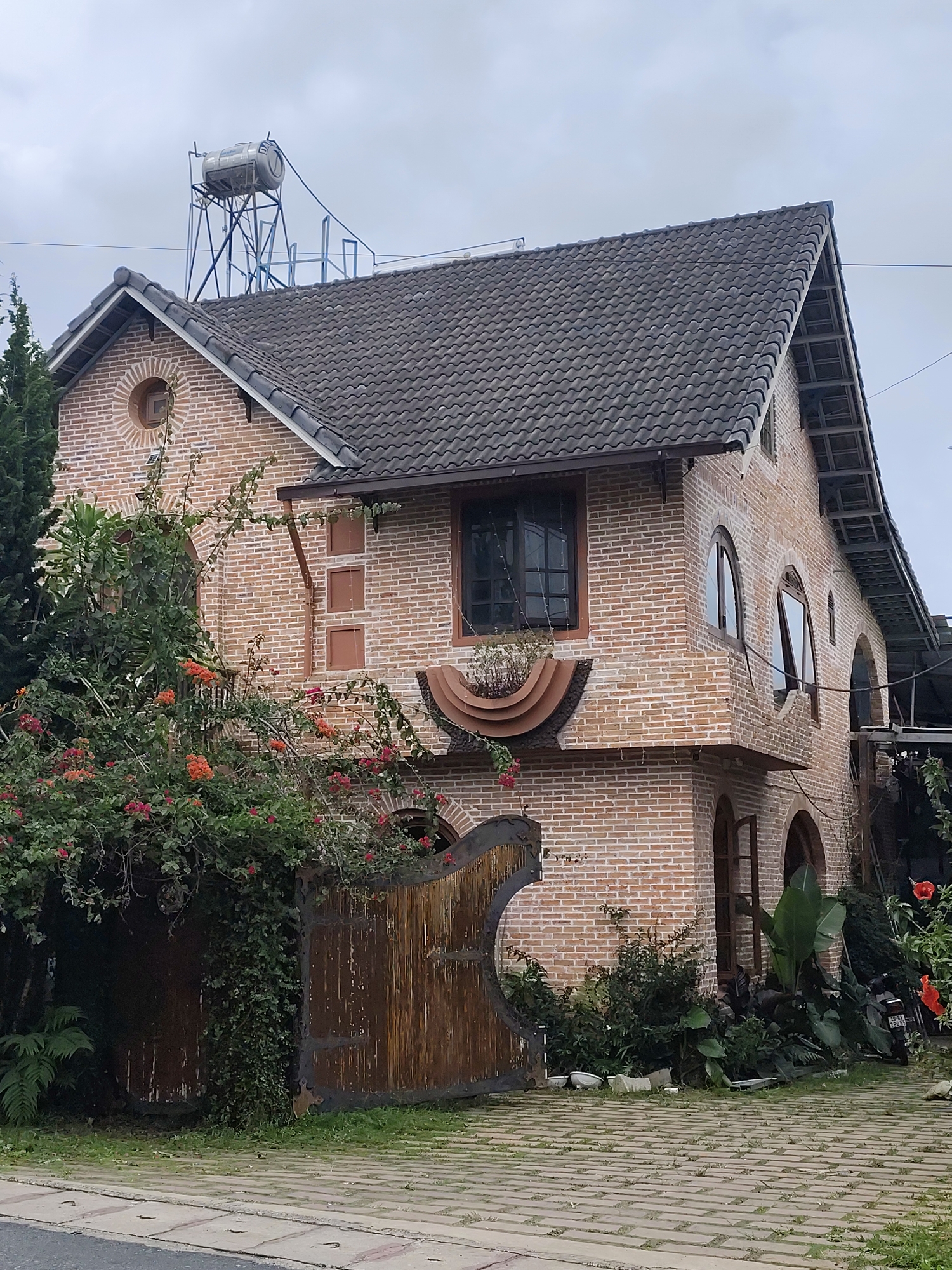 This image depicts a two-story brick house with a dark, textured tile roof. The architecture features arched windows and a circular window on the upper left gable. A large, rustic wooden gate with metal accents stands in front of the house, partially obscured by lush green foliage and vibrant orange and pink flowers. A water tank on a metal frame is visible on the rooftop. The house is situated on a paved pathway with visible grass growing between the stones, suggesting an outdoor or garden setting. The overall impression is of a charming, possibly older, residential or commercial property. The location is identified as Da Lat, Vietnam, a city known for its French colonial architecture and cooler climate, which would explain the prevalence of gardens and flowering plants.