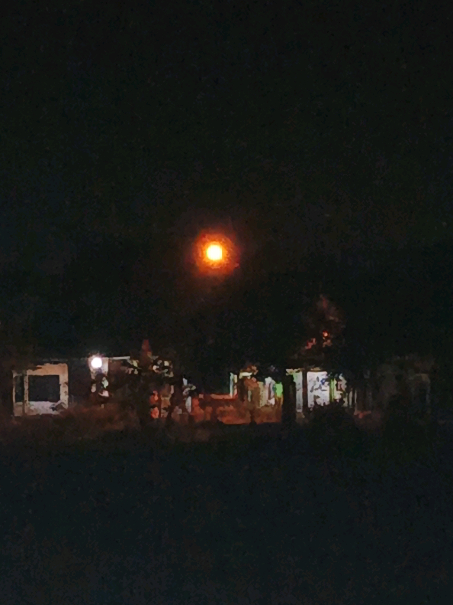 An outdoor night scene shows a dark background with a bright, orange, circular light source in the upper center. Below the light, a row of low-lying structures are partially illuminated by artificial lights. The structures appear to be made of concrete with dark openings. Trees and foliage are interspersed among the structures, their shapes indistinct in the low light. The foreground consists of dark, unlit ground. The overall impression is of a rural or semi-rural settlement at night, possibly in Ketegan, Indonesia, given the context of its location. The image's quality is low, with a grainy texture and limited detail.