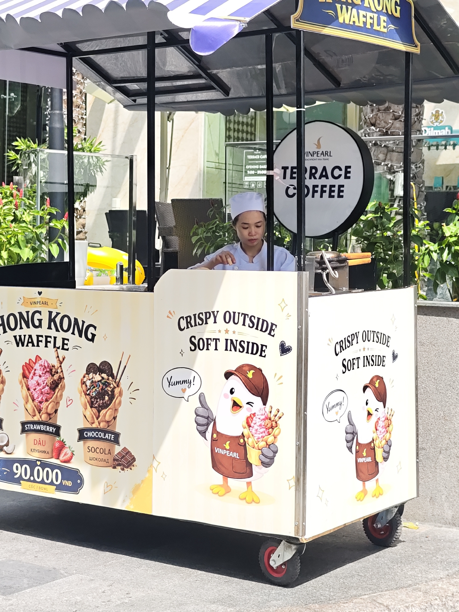 This is a medium-angle, eye-level shot of a food cart selling Hong Kong waffles. A woman wearing a white chef's hat and uniform stands behind the counter, appearing to be working. The cart is adorned with colorful graphics of waffles, cartoon chickens, and text promoting the product. The words "Hong Kong Waffle," "Crispy Outside Soft Inside," "Strawberry," and "Chocolate" are visible on the cart's display. A price of "90.000 VND" is also displayed. In the background, there are palm trees, suggesting a tropical or warm climate, and a sign for "Terrace Coffee" at "Vinpearl Nha Trang." The overall impression is a sunny day at an outdoor market or tourist area. The cart is on wheels, indicating it's a mobile food vendor. The cartoon chickens on the cart are depicted with happy expressions, and one is saying "Yummy!" suggesting a lighthearted and appealing atmosphere for customers. The Dilmah tea brand is also visible in the background.