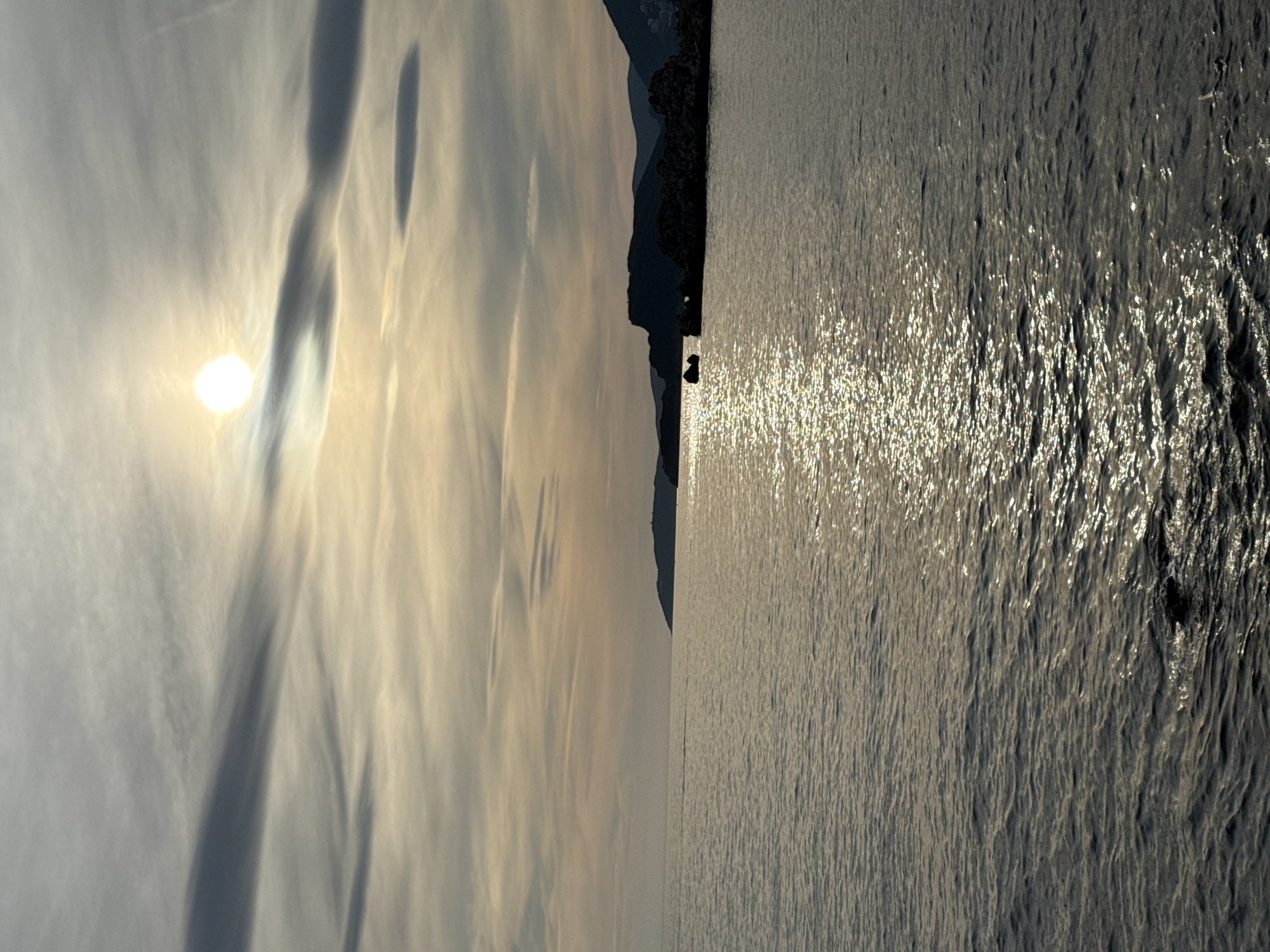 The image shows a view of the Adriatic Sea near Sveti Stefan, Montenegro. The sun is low in the sky, casting a bright reflection on the water. The sky is partly cloudy, with wispy clouds illuminated by the sun. In the distance, a silhouetted headland with rocky outcrops is visible, and further along the coast, the distinctive outline of Sveti Stefan island can be seen, though not in sharp focus due to distance and lighting. The sea is relatively calm with small ripples. There are no people or animals visible in the image. The overall impression is of a serene coastal landscape at dusk or dawn.