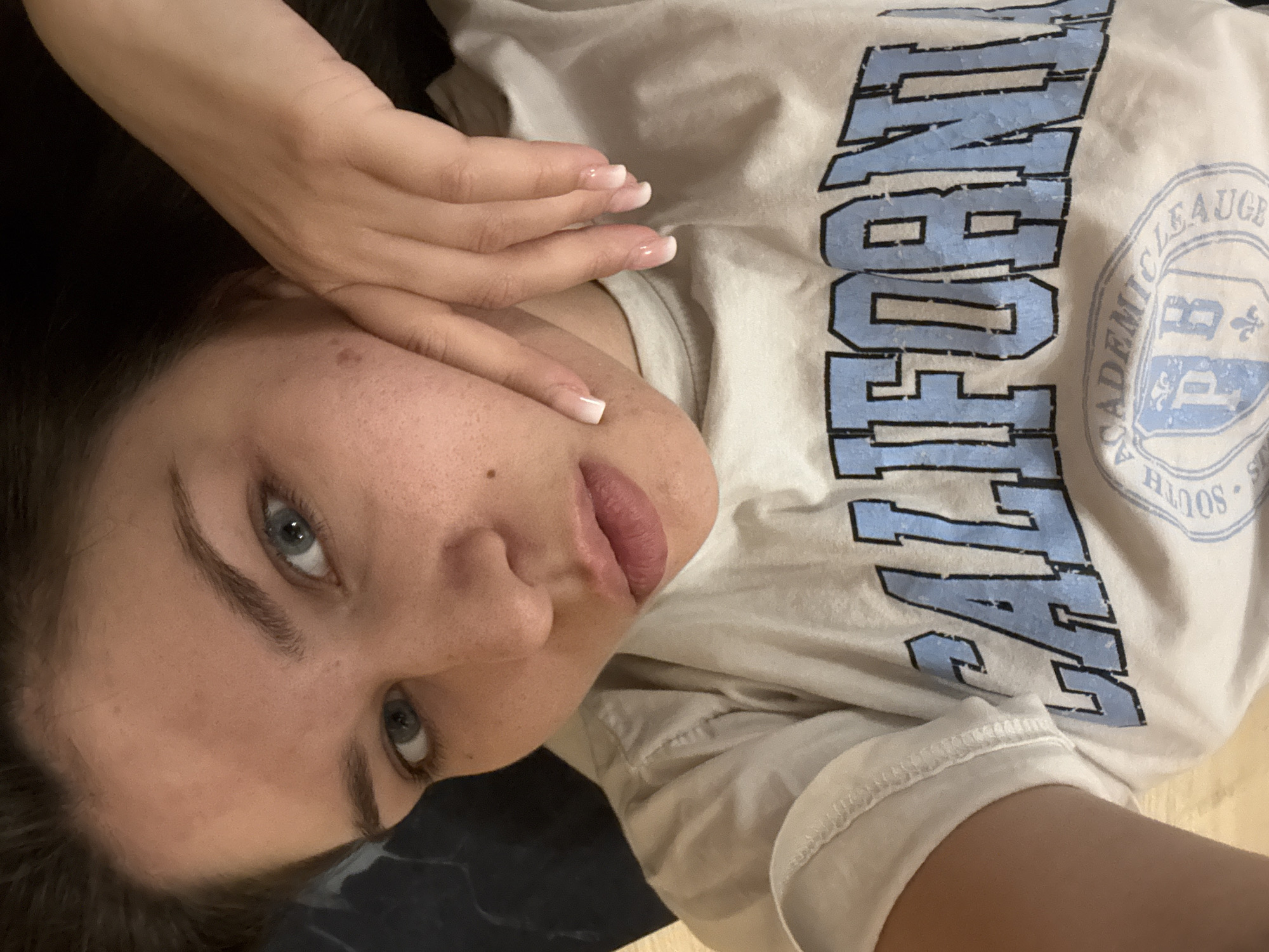 A woman lies on her back, her head tilted and resting on her left arm. Her face is angled towards the camera, with her blue eyes looking directly at the viewer. She has light skin with a smattering of freckles on her cheeks. Her lips are slightly parted, revealing a hint of her teeth. Her dark hair is spread out around her head.
She is wearing a light-colored t-shirt with large, light blue lettering across the chest. The text appears to be in a collegiate style. A circular emblem is also visible on the shirt, with text and a stylized "BP" inside. The t-shirt appears to be a casual, everyday garment.
Her left hand is raised and gently rests on her forehead, obscuring a portion of her hairline. Her fingers are elegantly posed, and her nails are neatly manicured with a French tip design. The background is out of focus, but suggests an indoor setting with a dark surface beneath her and a lighter surface to the right. The lighting appears to be soft and ambient, suggesting it could be daytime or an indoor light source. The overall impression is one of relaxed intimacy.