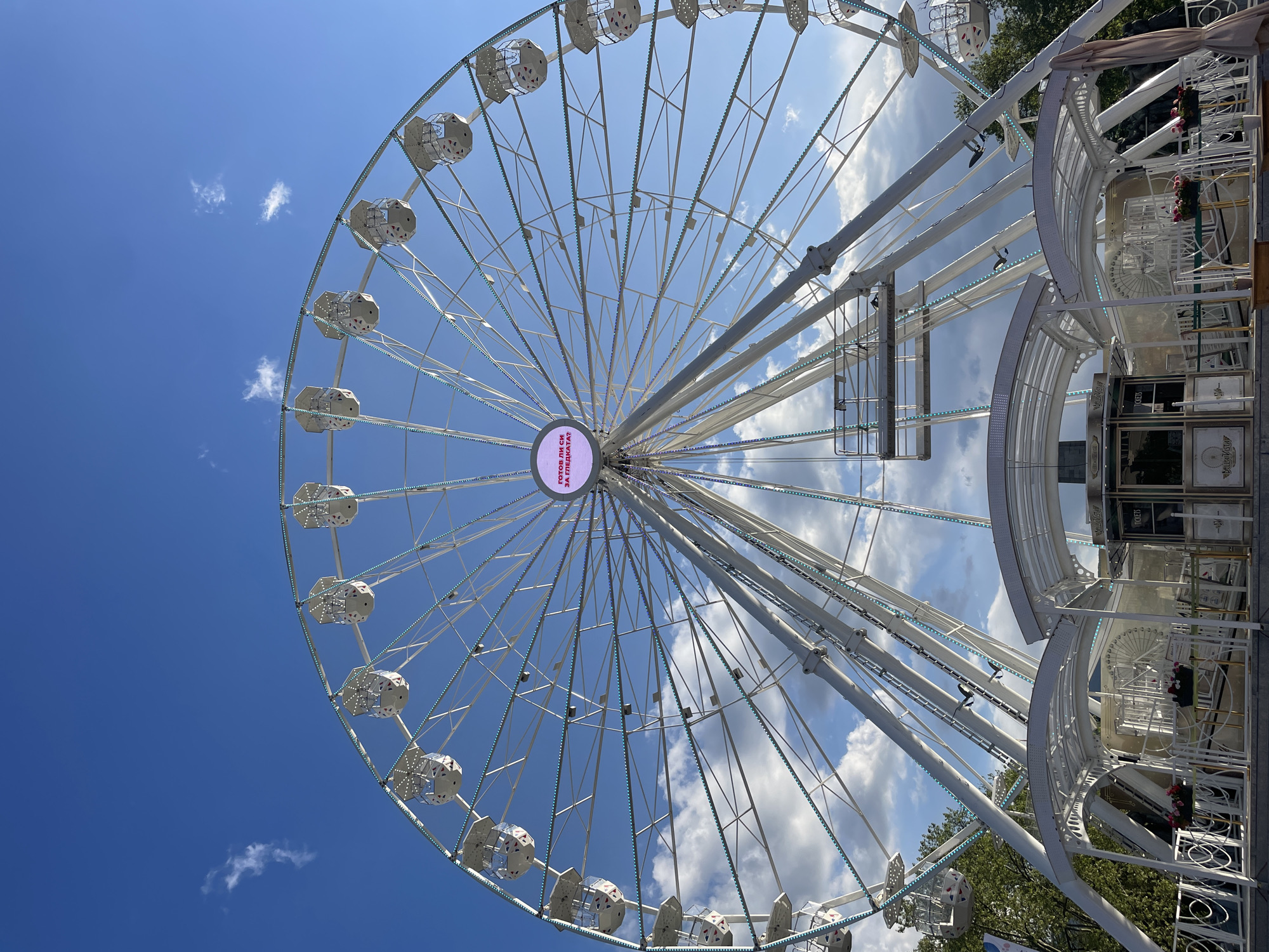 The image shows a Ferris wheel in Sofia, Bulgaria, under a blue sky with scattered clouds. The wheel is white, with numerous passenger cars. A circular display at the center of the wheel is lit up. Adjacent to the wheel is a structure that appears to be the entrance or a support structure for the wheel. Trees are visible on the periphery.