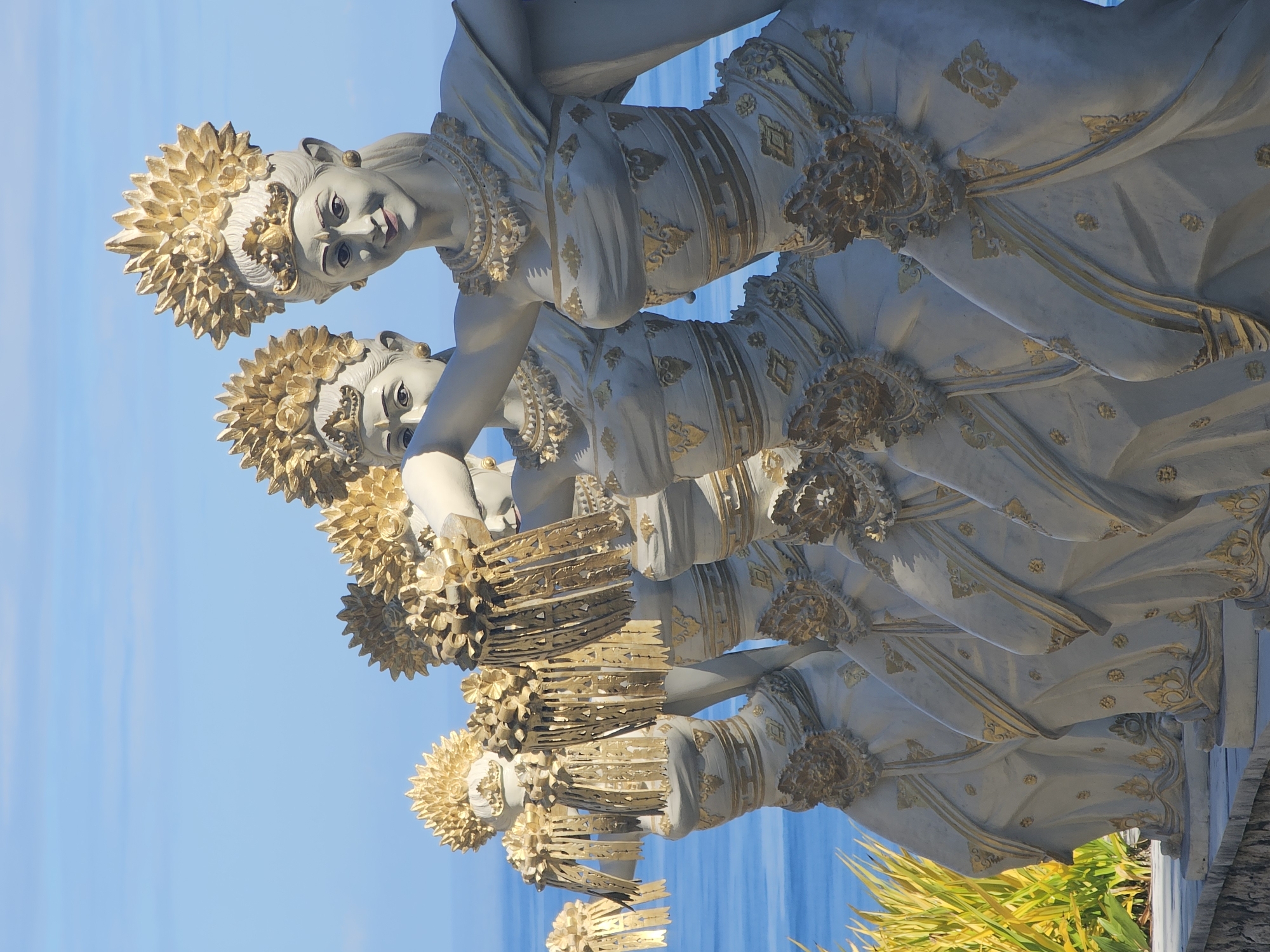 The image shows a series of four identical statues of Balinese dancers, arrayed in a row. The statues are predominantly white, with gold accents on their headdresses, necklaces, dresses, and ornaments. The dancers have serene expressions, looking downwards and slightly to the left.
The backdrop consists of a clear blue sky and a narrow strip of the ocean. The statues are located in the city of Kuta Selatan, Indonesia. Green and yellow plants are visible at the bottom right corner of the frame, near the statues.