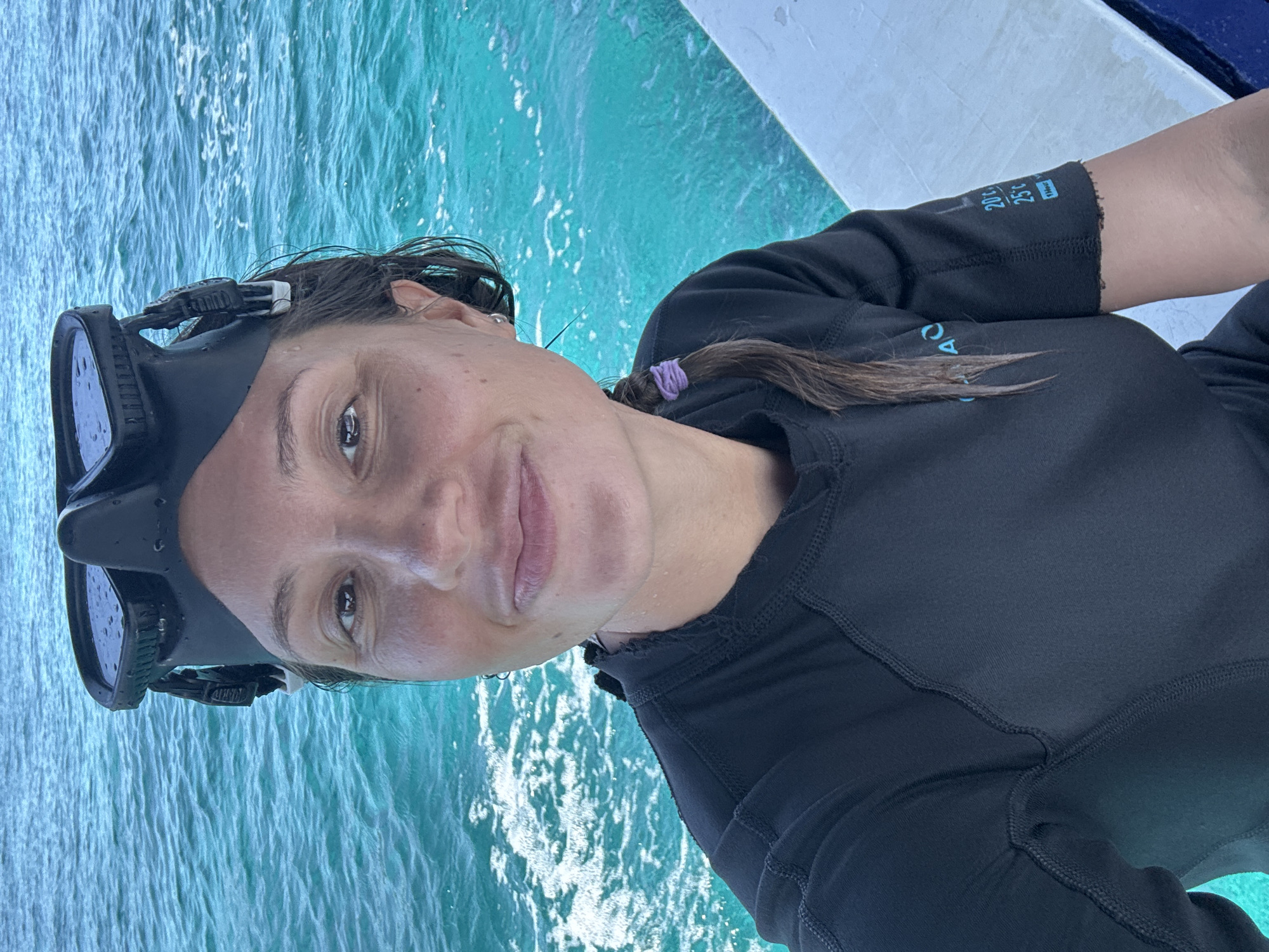 A woman is positioned in front of a turquoise body of water, likely the ocean, as indicated by the water's color and texture. She wears a black diving mask on her forehead, and a black, long-sleeved rash guard, which has markings on the right sleeve including temperatures in Celsius. She has a dark braid with a purple hair tie, pulled to her right. The location is Unknown, Indonesia.
