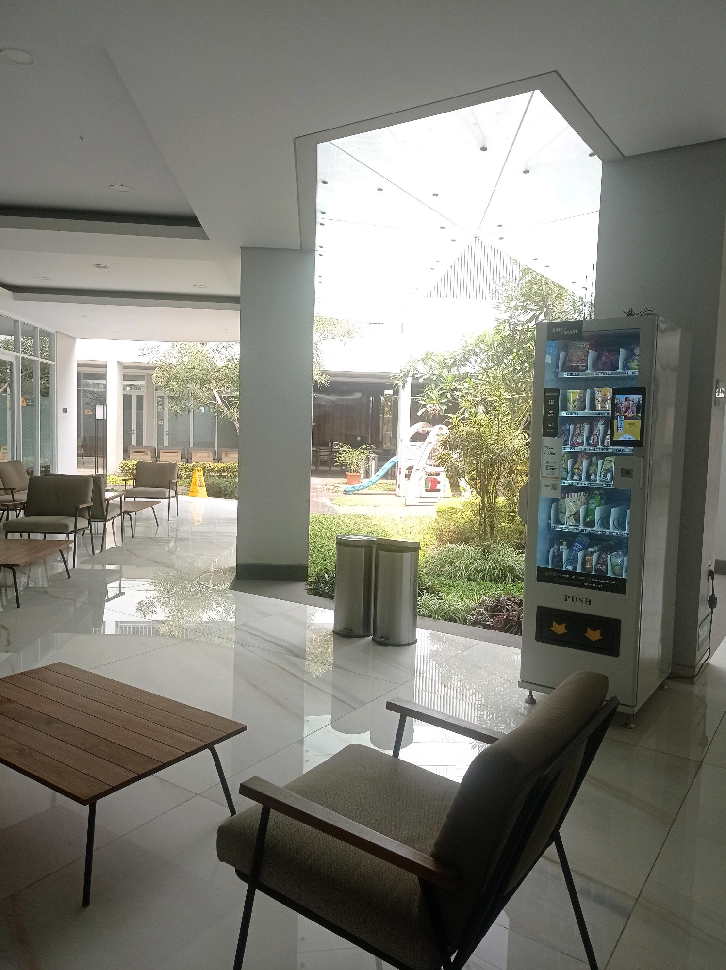 The image depicts an indoor waiting area with a modern design, located in a building in Bandung City, Indonesia. The area is characterized by a glossy white tiled floor that reflects the abundant natural light entering from a large, angled skylight and floor-to-ceiling windows. To the right, a white vending machine is prominently placed, filled with various beverages and snacks. Next to the vending machine are two cylindrical silver trash bins. In the foreground, a wooden coffee table with black metal legs sits next to a tan upholstered armchair with a dark metal frame, facing away from the camera. Further into the space, more seating arrangements are visible, including armchairs and small tables, arranged around a central outdoor courtyard. The courtyard contains lush green plants, a small playground structure with a blue slide, and more seating. The overall impression is one of a clean, well-lit, and comfortable space designed for waiting or relaxation.