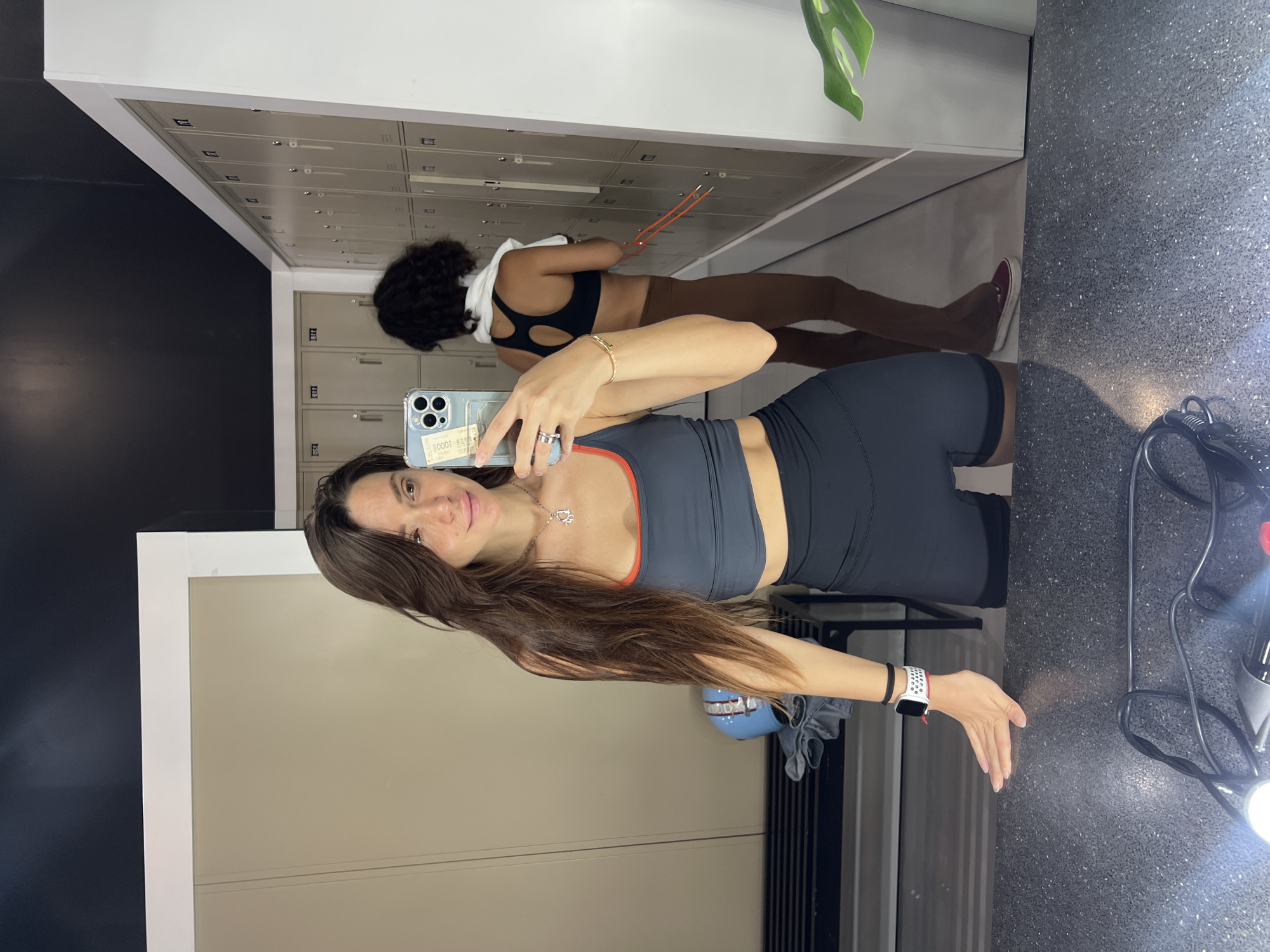 A woman takes a mirror selfie in a gym. The woman is wearing a gray sports bra and dark gray shorts. She has long, brown hair. Another woman is visible in the mirror, wearing a black sports bra, brown leggings, and red shoes. They are in front of gray metal lockers. A white towel hangs over the second woman's shoulder. A plant is visible in the upper right corner of the mirror. A black, speckled countertop and workout equipment are visible in the lower right corner.