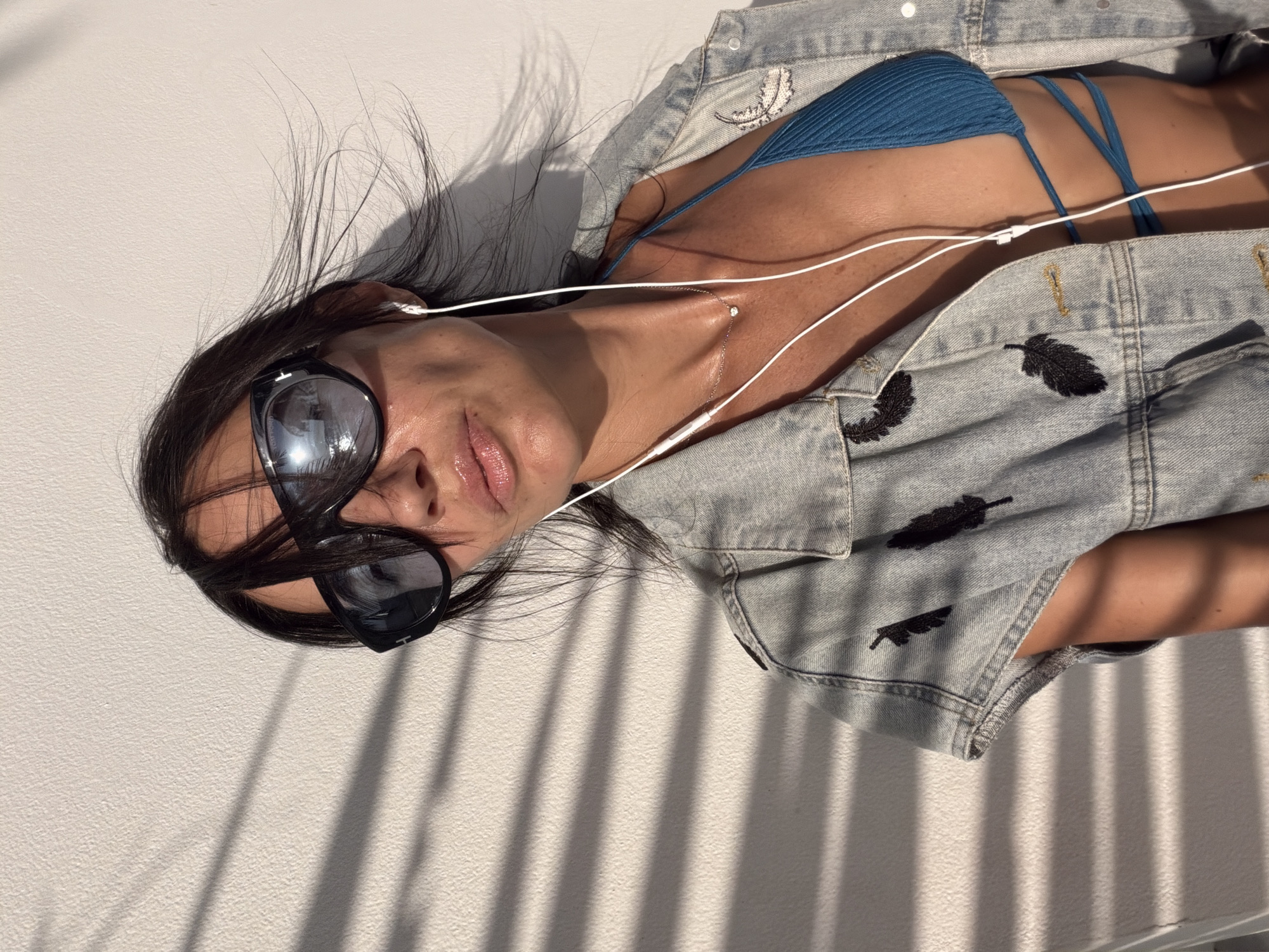A woman with dark hair and sunglasses poses against a textured, white wall. She is wearing a blue bikini top and a denim jacket adorned with black feather patches. White earbuds are in her ears, and the cord trails down her chest. Light casts horizontal shadows on the wall and her body. The scene appears to be an outdoor setting, possibly a balcony or patio, given the natural light and the presence of shadows. The woman's pose is relaxed, with her head tilted slightly. The overall impression is one of casual confidence in a sunny, outdoor environment.