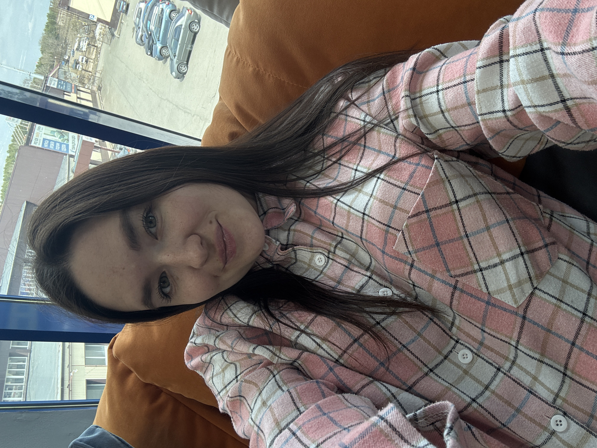 A young woman with dark hair and fair skin is taking a selfie. She is wearing a pink and white plaid shirt and appears to be sitting on an orange cushion.
Through a window, a parking lot with several cars is visible. The building behind the parking lot has signs in Russian, suggesting the location is likely in Russia. Based on the signs and available details, the location is in Chaikovsky, Russia.
