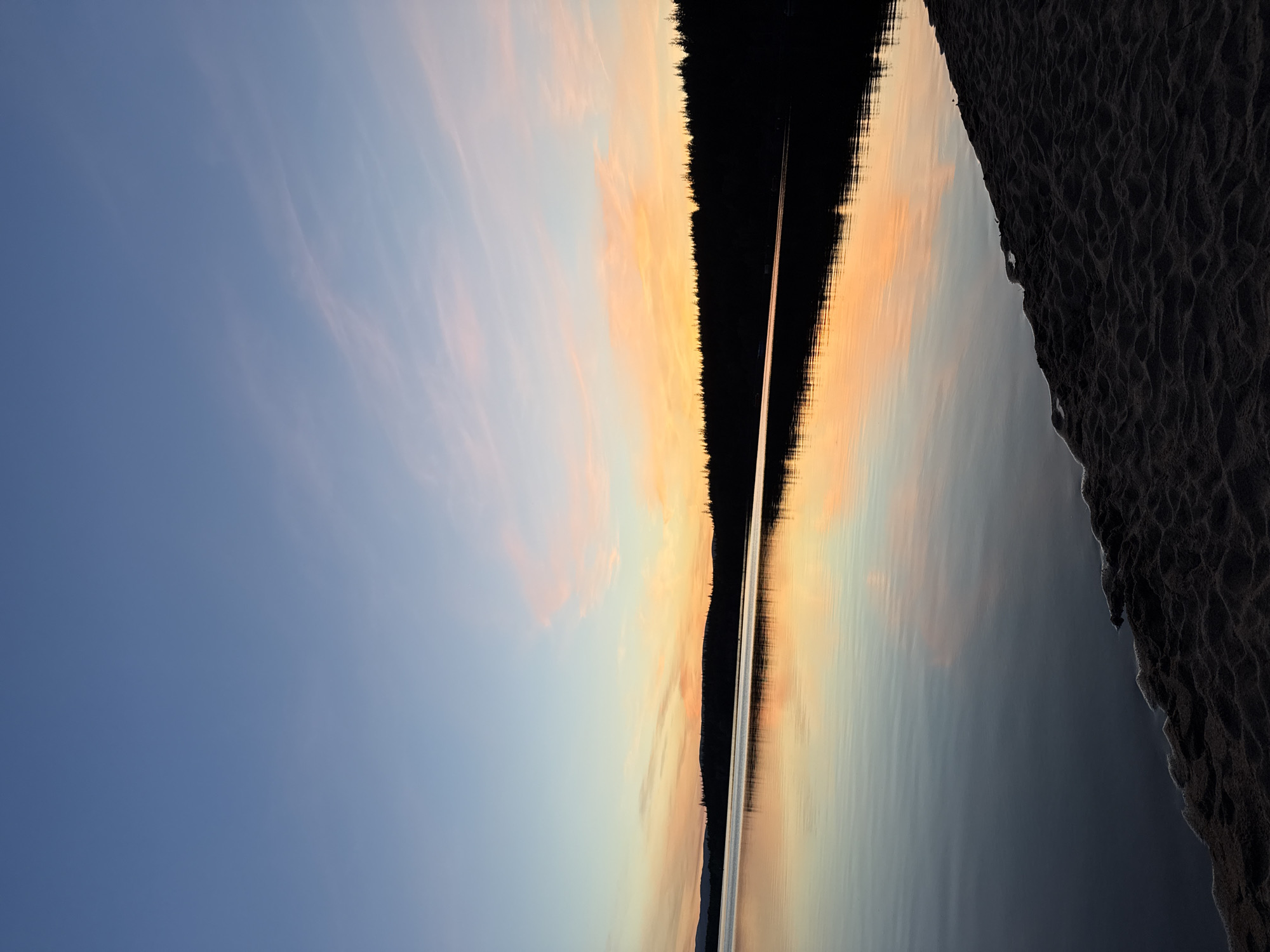 The image displays a tranquil natural landscape captured during either sunrise or sunset. The scene is composed of a sky, a distant treeline, a body of water, and a foreground shore.
The upper left portion of the image features a clear, deep blue sky, which gradually transitions to soft pastel shades of pink and orange towards the center. Subtle, wispy clouds are faintly visible against the blue and lighter hues.
A prominent, dark silhouette of a treeline stretches horizontally across the middle of the frame, delineating the horizon. This dense, dark band of trees suggests a distant forested shore or island.
Below the treeline, a calm body of water, likely a lake, perfectly mirrors the sky's colors, reflecting the gentle blues, oranges, and pinks. A distinct, narrow streak of bright light extends horizontally across the water's surface, indicating the reflection of the sun or a light source. The water appears smooth and still, enhancing its reflective quality.
In the bottom right corner, a dark, textured area occupies the foreground, resembling sand or earth with subtle undulations, marking the immediate shoreline.
The overall impression is one of peace and serenity. No human subjects, man-made objects, or visible text are present, emphasizing the undisturbed natural beauty of the moment. The soft lighting and calm water suggest a quiet time of day.