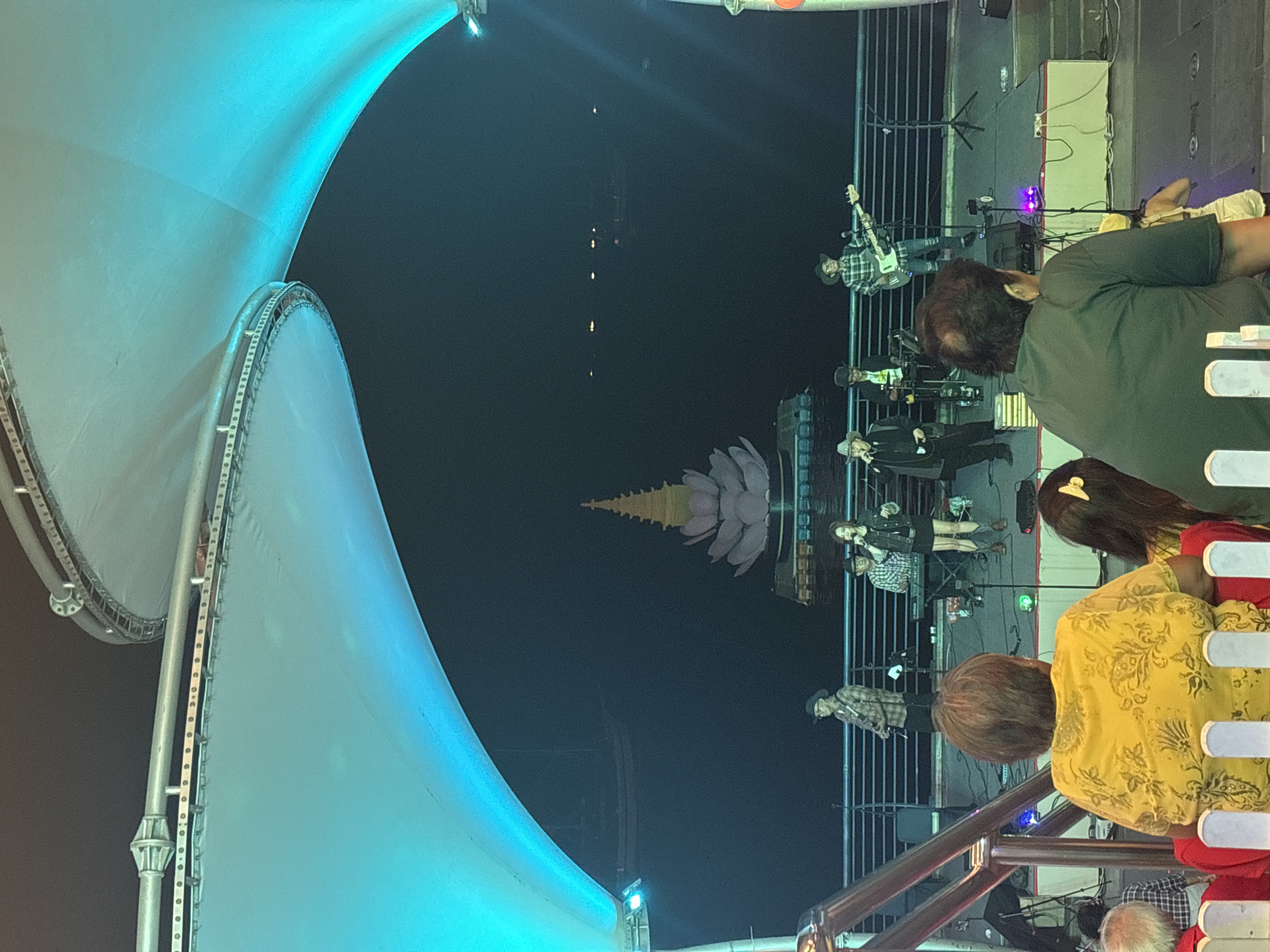 The image shows an outdoor concert at night, likely in the city of North Jakarta, Indonesia, as the Bundaran HI Lotus Monument is visible in the background. A band of about five members is performing on a stage behind a railing. Several spectators are watching the performance from behind the railing. The scene is lit with stage lights and an illuminated structure, possibly a tent or canopy, dominates the left side of the image.