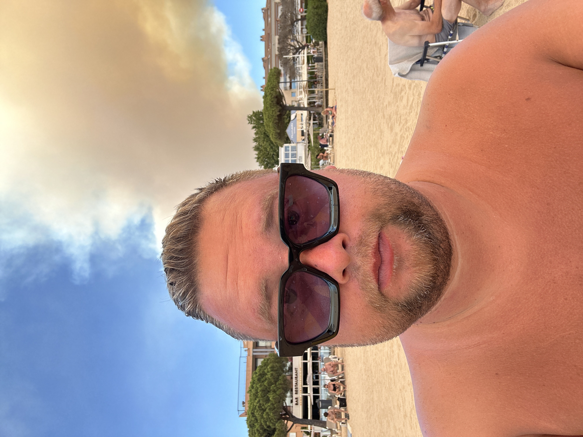 The image is a selfie taken at the beach in Sant Feliu de Guíxols, Spain. A man wearing sunglasses is in the foreground, facing the camera. In the background, there is a sandy beach with several people relaxing on lounge chairs. Beyond the beach, there are buildings, trees, and a cloudy sky with a yellowish hue, possibly indicating smoke or dust. A sign on one of the buildings reads "BAR RESTAURANT."