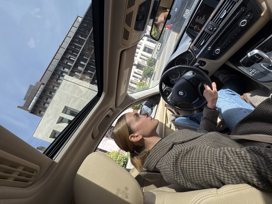 The image captures the interior of a BMW vehicle, specifically from the front passenger's perspective looking towards the driver and the exterior. A woman with light-colored hair styled in a low ponytail occupies the driver's seat, wearing a brown and tan patterned long coat over a dark top and light blue jeans. Her left hand rests on the black multi-function steering wheel. Her head is turned to her right, gazing forward. Her reflection is partially visible in the rearview mirror. The car's interior features beige upholstery on the seats and ceiling, with black elements on the dashboard and center console, which includes an automatic shifter. An open panoramic sunroof reveals a clear, bright blue sky.
The scene is set on a sunny day in an urban environment in Tbilisi, Georgia, confirmed by a Georgian license plate "PZ-152-FL" visible on a black vehicle reflected in the rearview mirror. Outside the vehicle, modern multi-story buildings are visible. A prominent light-colored building with large, recessed window sections stands to the left. Behind it and to the right, a taller, darker building with a distinct linear, possibly metallic or dark stone, facade is seen against the sky. Green foliage from trees or bushes lines the street visible through the windshield and rearview mirror.