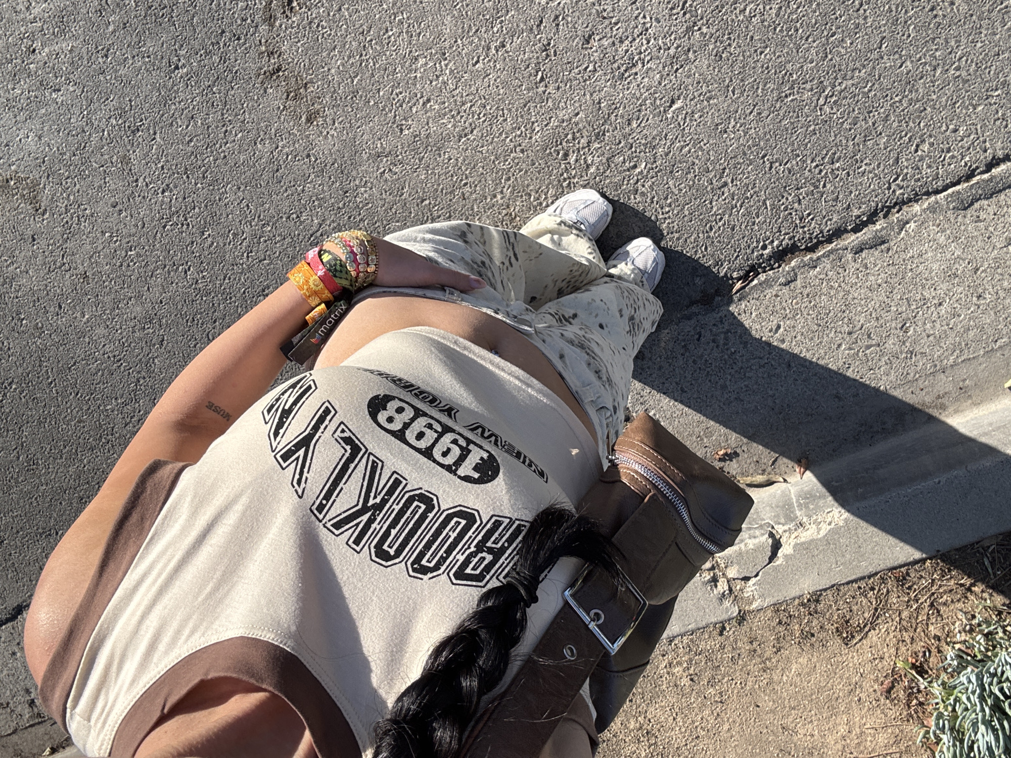The image shows a person standing on concrete, likely a sidewalk or road, with a partial view of their outfit and surroundings. The person is wearing white sneakers, beige pants with a splatter pattern, and a beige tank top with brown trim and the word "BROOKLYN" printed on it. They have several bracelets on their left wrist and a brown bag with a silver buckle and zipper. Their hair is styled in a braid. The concrete surface shows signs of wear and there is a curb and some ground cover visible in the background. The location is Newport Beach, United States of America.