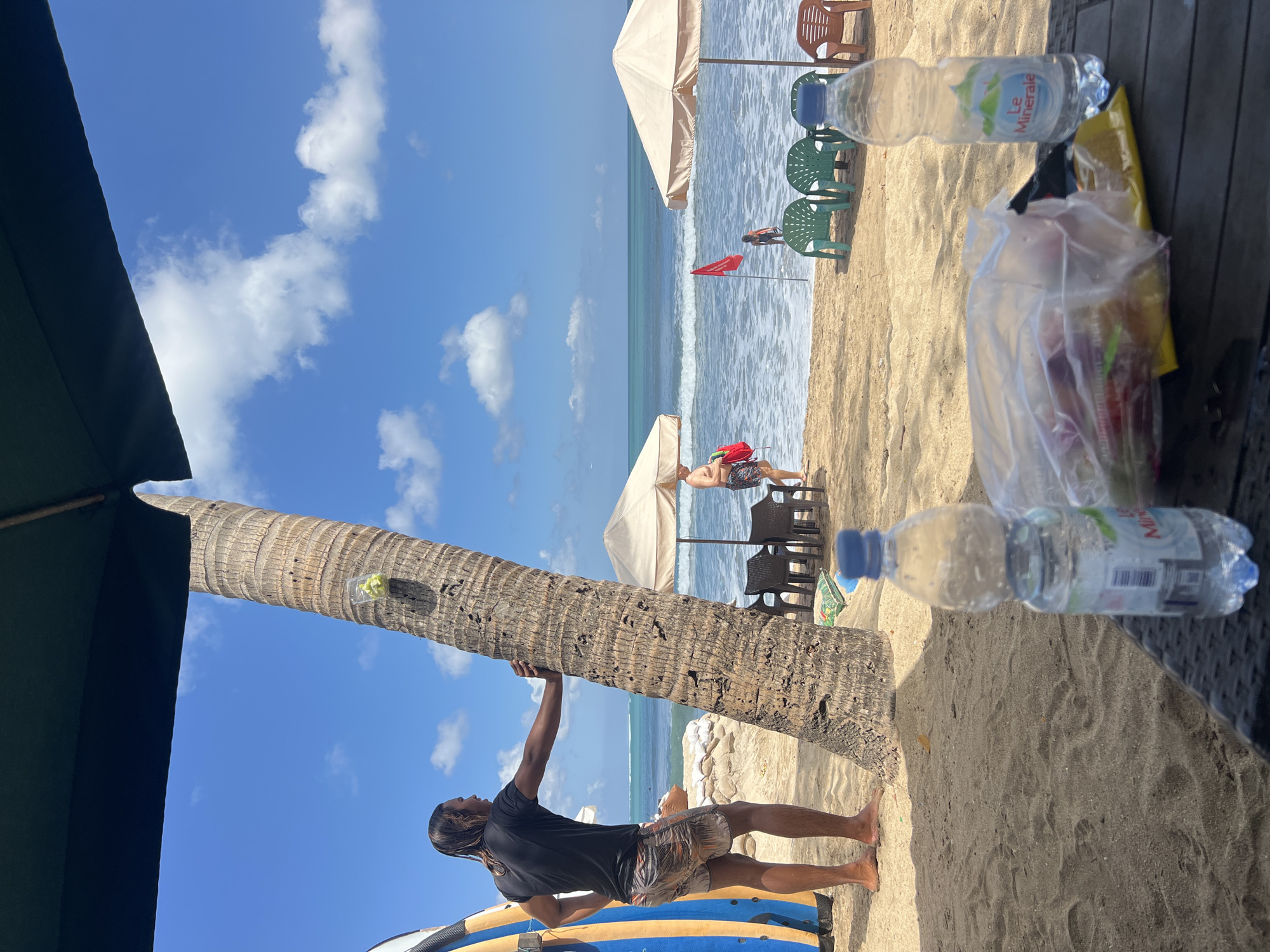 This image depicts a beach scene in Kuta, Indonesia, on a sunny day with a clear blue sky and scattered white clouds. In the foreground, a table holds a bottle of "Le Minerale" water and a clear plastic bag filled with various items, possibly food. Beyond the table, a sandy beach stretches towards the ocean, where gentle waves are breaking.
Several beach umbrellas and chairs are set up on the sand, indicating a leisure area. A prominent palm tree stands near the center of the frame, with a man in a black t-shirt and patterned shorts leaning against it, looking out at the sea. To his left, a blue and yellow surfboard is propped up. In the background, other beachgoers are visible near the water, including a man in patterned shorts carrying a red bag. A red flag is planted in the sand near the water's edge. The overall impression is one of a relaxed tropical beach environment.