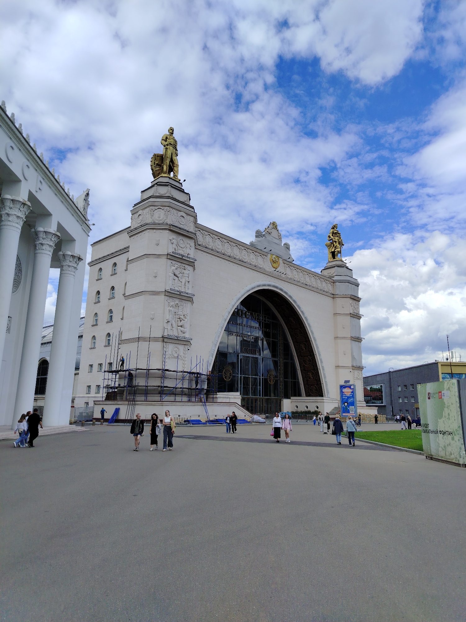The image depicts a view of the Pavilion of Agriculture at VDNKh (Exhibition of Achievements of National Economy) in Moscow, Russia, on a cloudy day. The main structure is a large, cream-colored building with classical architectural elements, including columns on the left side and decorative sculptures atop the facade. A prominent arched entrance features a large glass window. Scaffolding is visible on the facade of the main building, indicating ongoing construction or renovation work. Several people are walking on the paved area in front of the pavilion. The sky is partly cloudy with patches of blue sky visible. In the background, other buildings and a green grassy area can be seen. A large sign with Russian text is also visible on the right side of the image.