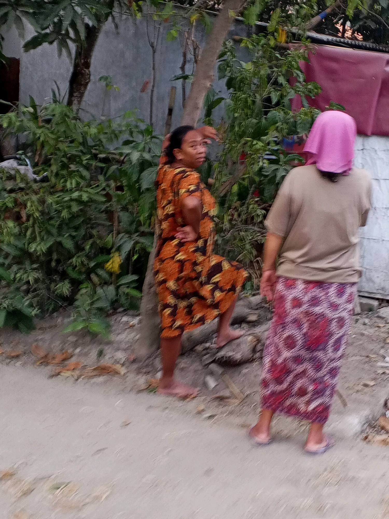 Two individuals are standing near some foliage and a wall in what appears to be a residential area. The person on the left is facing away from the camera, with their left hand on their hip and their right hand touching a tree trunk. They are wearing an orange and brown patterned dress. The person on the right is also facing away from the camera, wearing a light brown t-shirt and a long skirt with a red and purple pattern. They have a pink head covering. The ground is dirt and sand, with some scattered leaves and debris. A pink tarp is visible in the background. The scene is outdoors, with trees and plants present. The overall lighting suggests it is daytime, possibly late afternoon. The location is identified as Kepandean, Indonesia.
