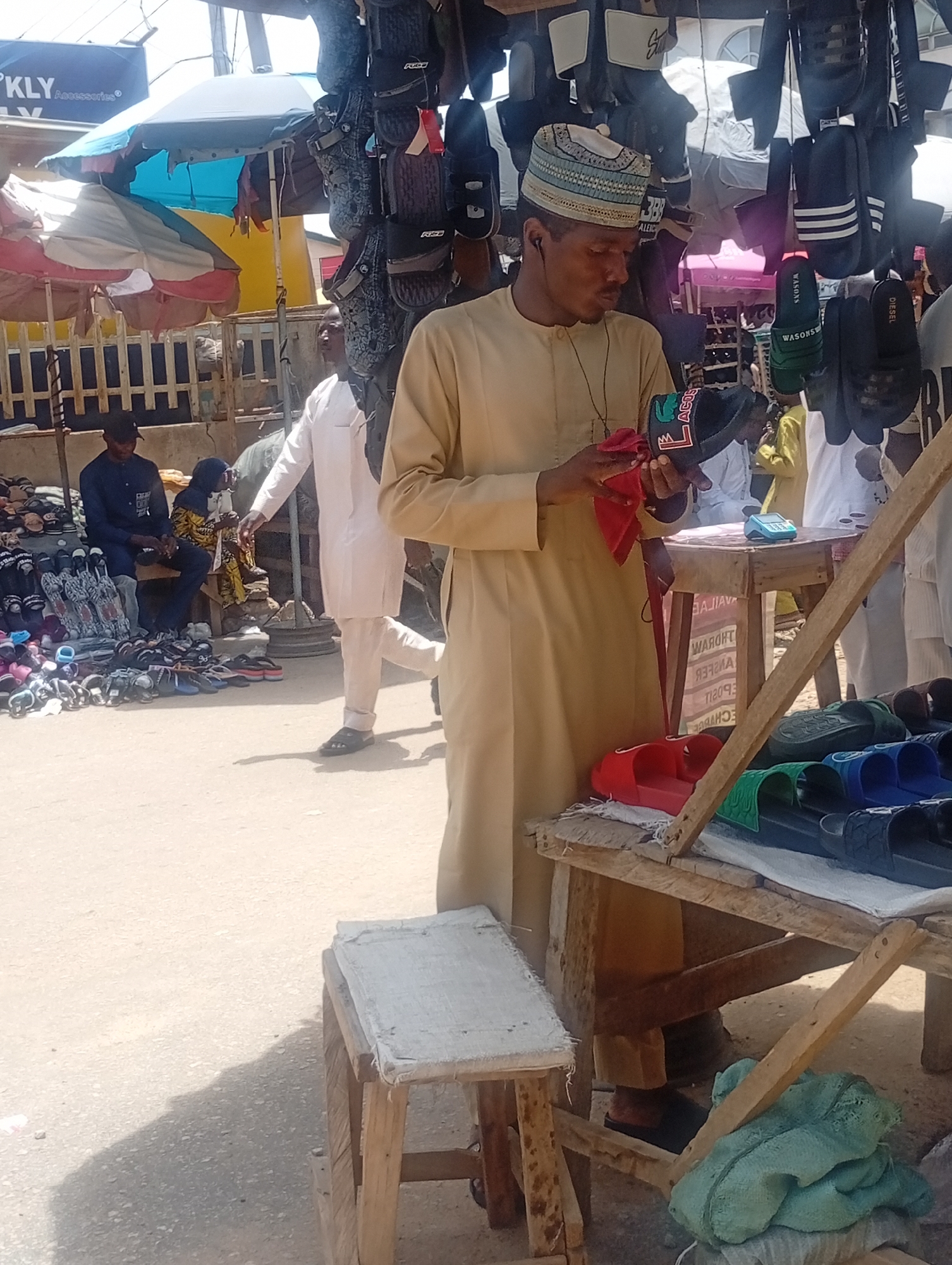 A street-level view of an outdoor market in Maiburiji, Nigeria, with a man dressed in a light brown robe and a patterned cap standing at a wooden display table cleaning a black sandal with a red cloth. Hanging above him are numerous pairs of sandals. Another display table to his right shows an assortment of colorful sandals. In the background, several people are visible, including two men sitting near a larger display of sandals and a man in a white robe walking away from the camera. Umbrellas provide shade overhead.