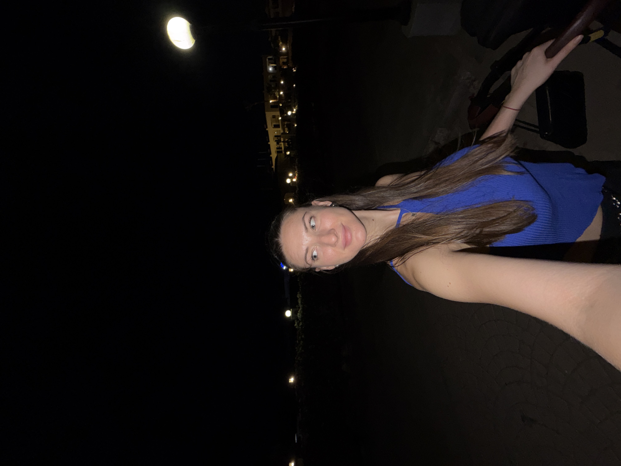 A woman with long brown hair, wearing a blue ribbed halter top and black pants, stands in the foreground and takes a selfie. She is outdoors at night, with a streetlamp to her right and buildings illuminated in the distance. The ground appears to be paved. The background shows a dark, unlit expanse with scattered lights from buildings. The general impression is a nighttime scene on a walkway, possibly near a resort or residential area in Sharm El Sheikh, Egypt, given the warm climate suggested by the woman's attire and the nighttime setting.