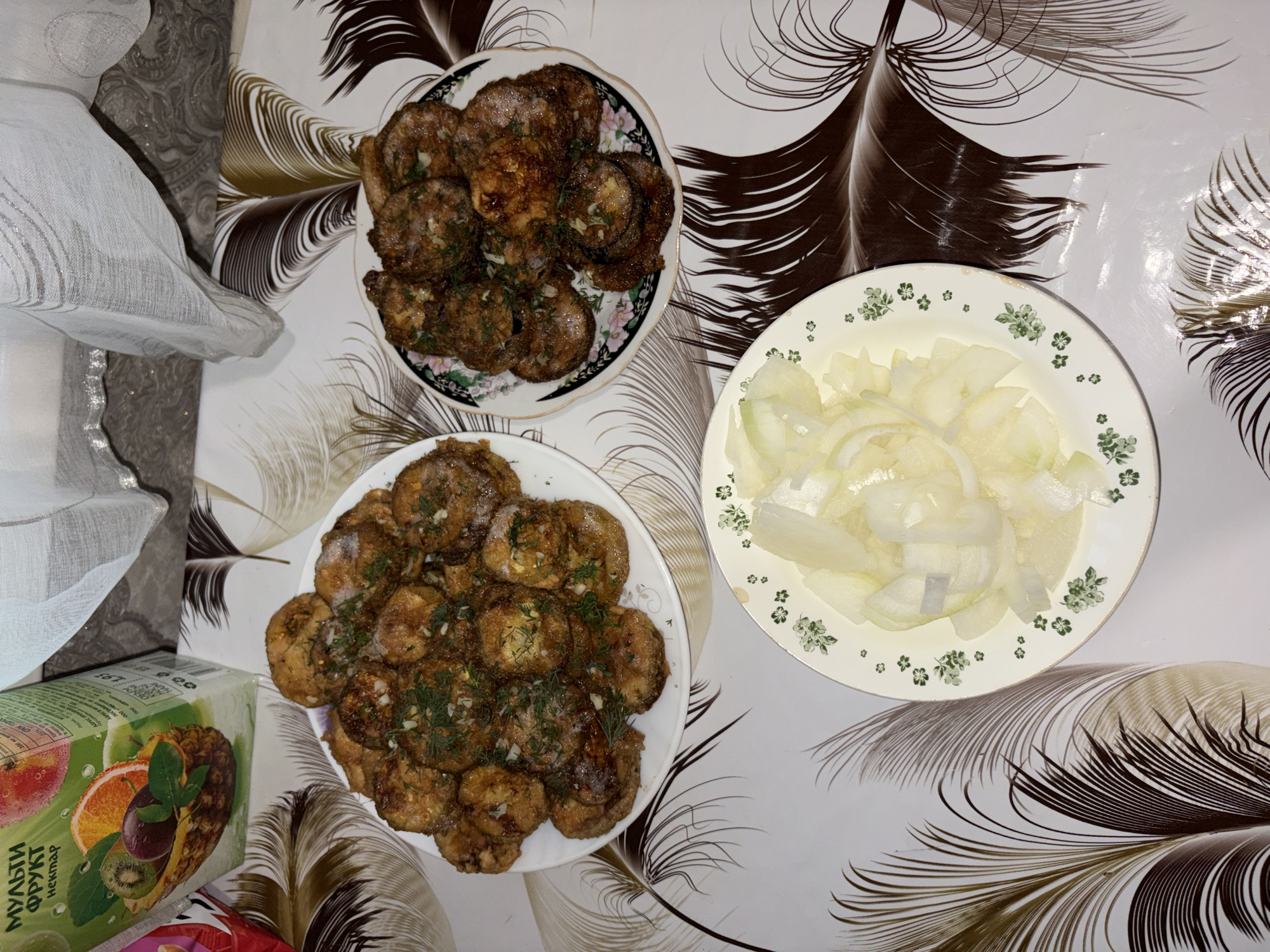 The image shows a table with food and a juice box. There are three plates of food. Two plates contain fried round slices of what appear to be zucchini or eggplant, seasoned with herbs and garlic. One plate is filled with sliced onions. A juice box with "Мульти фрукт нектар" printed on it is visible in the upper left corner. The table is covered with a tablecloth featuring a brown and white feather-like pattern. The scene appears to be indoors. No specific location in Tambov, Russia can be determined from the image.