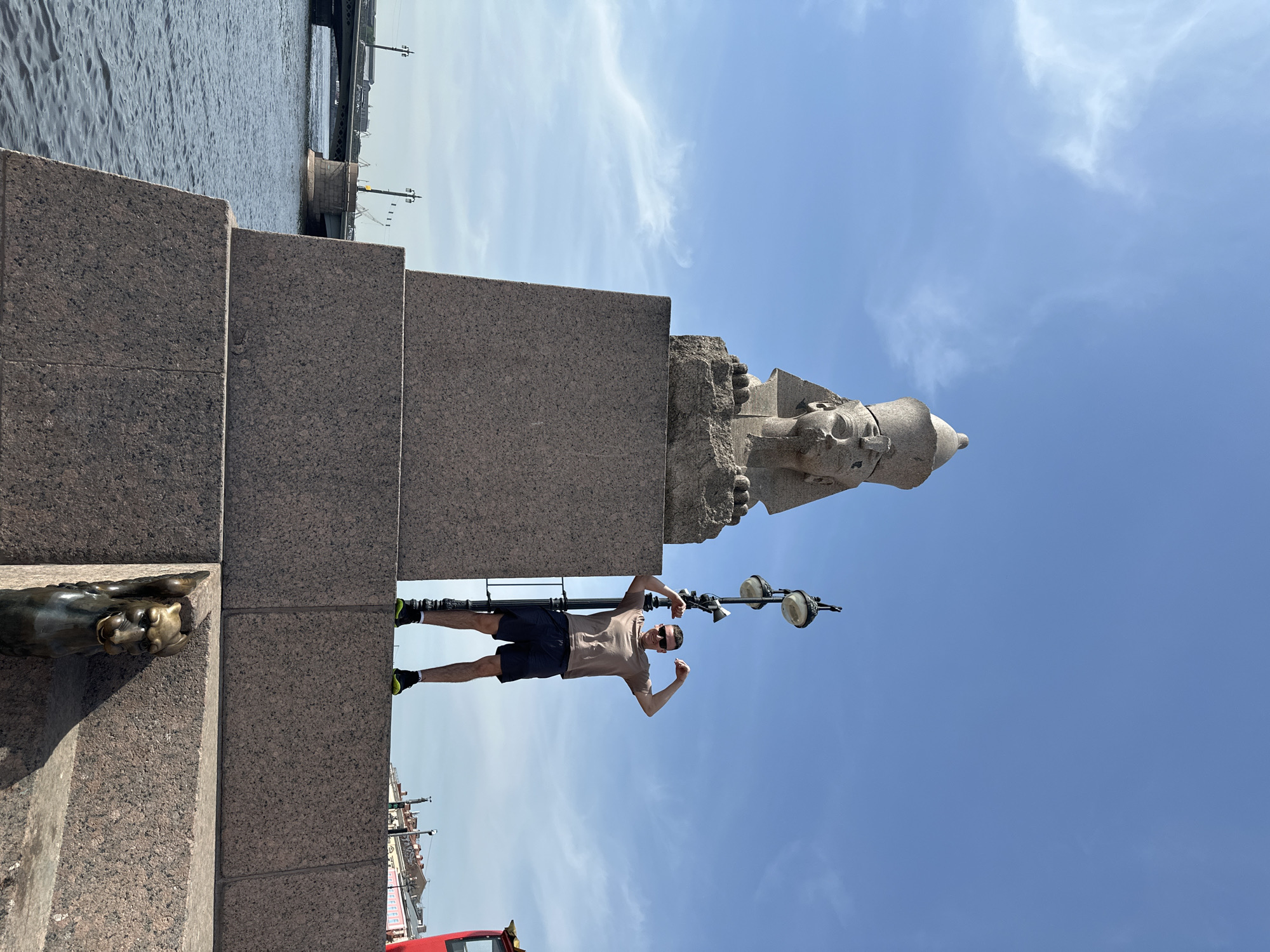 A man stands beside a Sphinx statue and a lamp post on a granite structure, under a blue sky with white clouds. The location is a riverside embankment in St. Petersburg, Russia, identifiable by the sphinx statues. The man is wearing shorts, a t-shirt, and sunglasses, and is posing by flexing his arms. A stone lion statue is partially visible on the lower left. A red vehicle is visible in the bottom portion. A bridge is seen over water in the background.