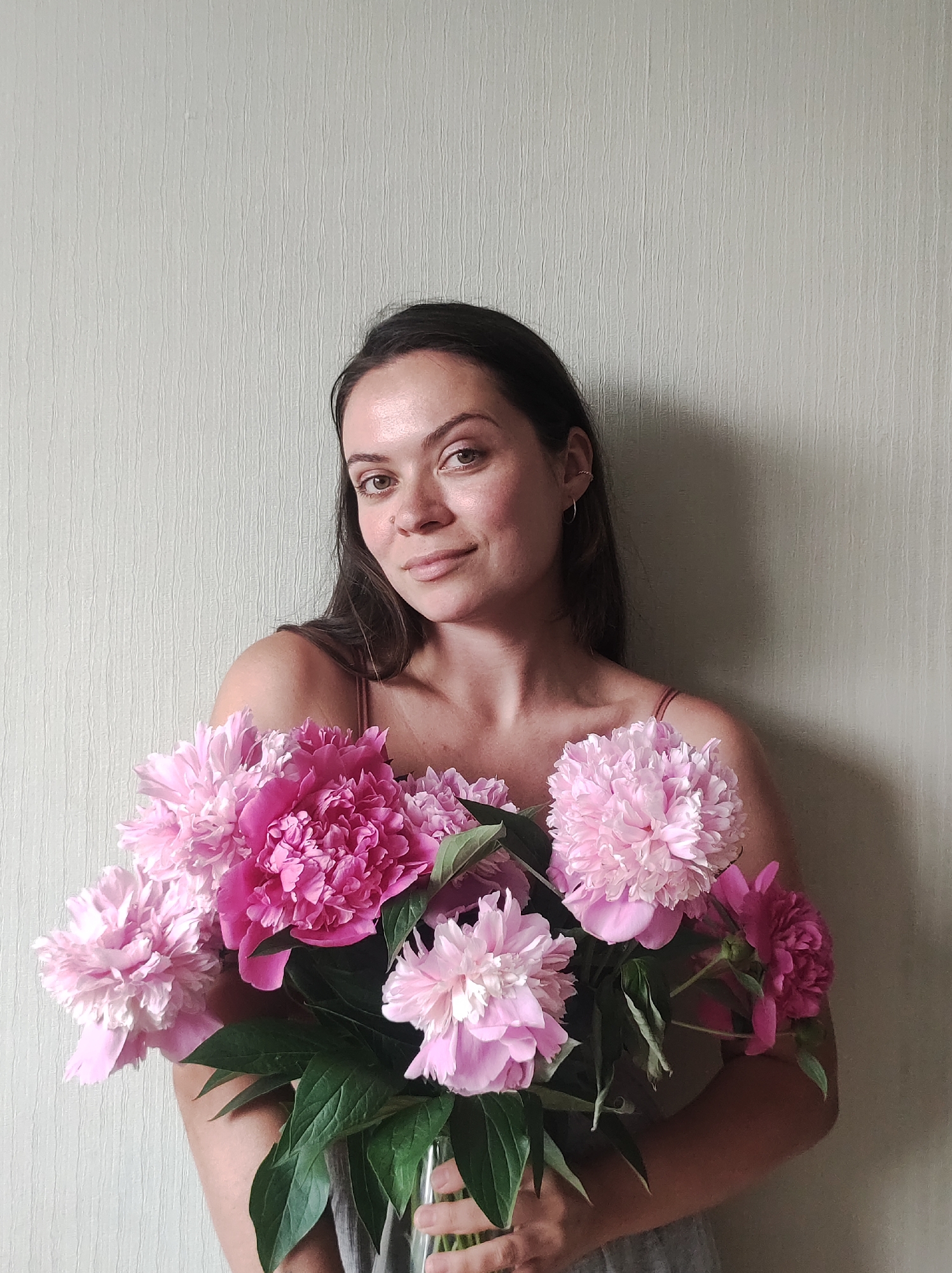 A young woman with long, dark hair and fair skin is holding a bouquet of pink peonies in front of a textured, pale green wall. She is wearing a light-colored camisole. The background appears to be an interior wall. The image does not contain any specific landmarks or clues that would indicate its location in the city of Chaikovsky, Russia.