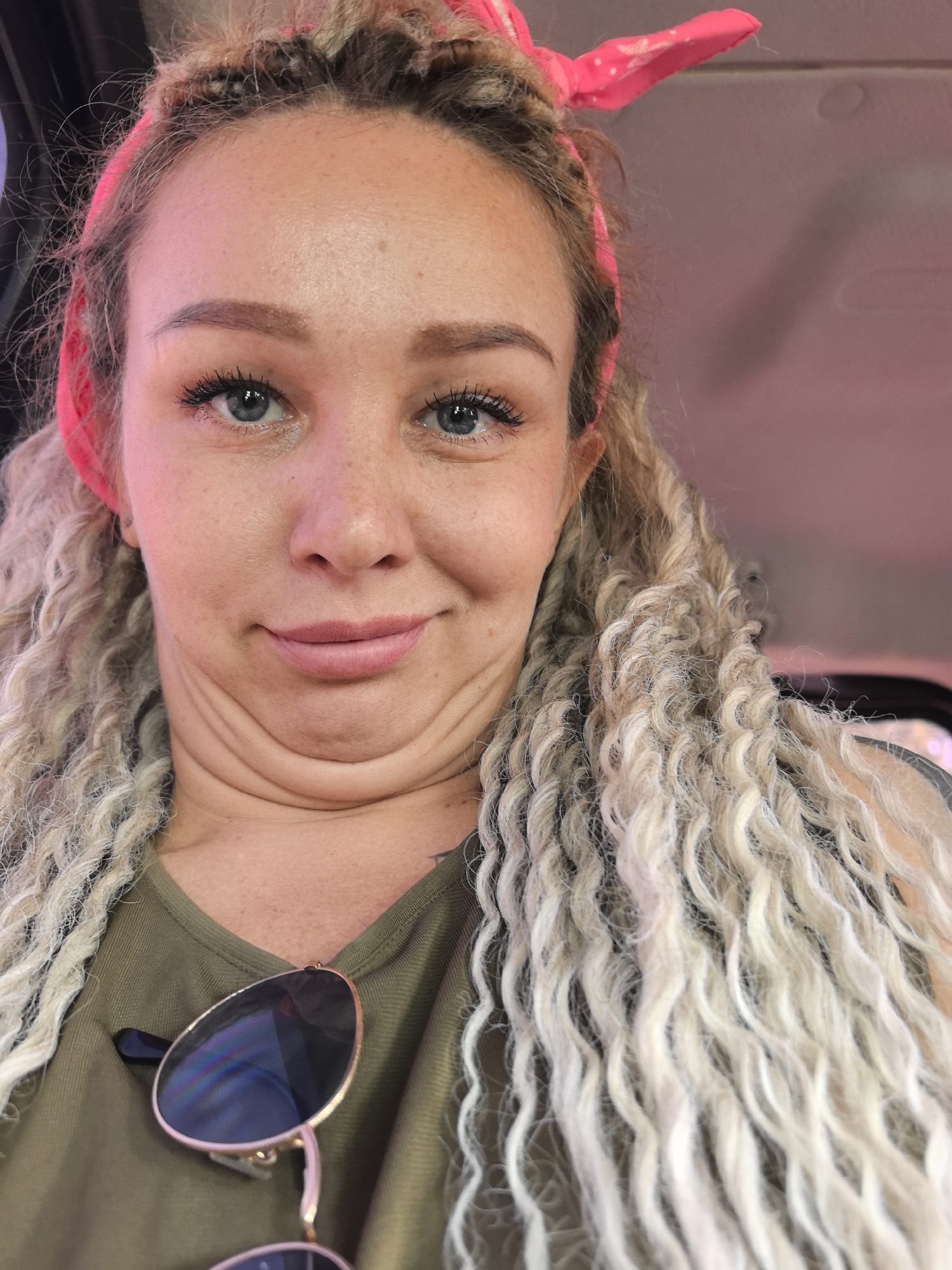 A selfie of a fair-skinned woman with blond, curly hair wearing a pink headband. She has blue eyes and is wearing a green v-neck shirt with sunglasses attached. The background appears to be inside a vehicle with a pink interior. There's no explicit indication that this image was taken in Chaikovsky, Russia.