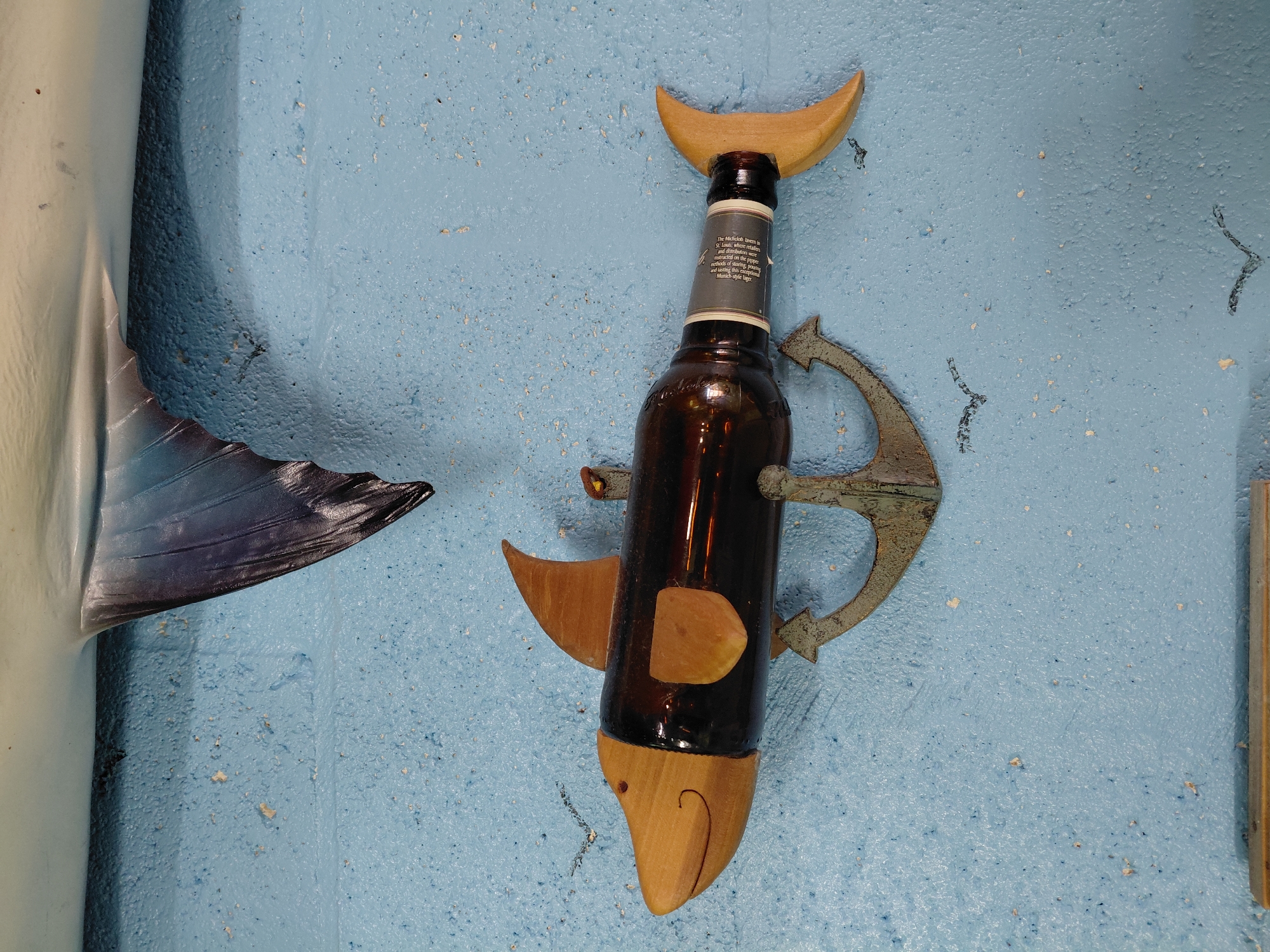Here is a detailed description of the image:
The image shows a wall decoration in the shape of a fish, composed of a brown beer bottle with wooden attachments. A wooden crescent moon shape is affixed at the top of the bottle, forming the dorsal fin. Wooden pectoral fins are attached to the sides, and a curved wooden piece acts as the tail and lower part of the fish's head, with a small hole representing the eye and a curved line showing the mouth. An old, weathered anchor is attached to the middle of the bottle. To the left of the fish is a partial image of a printed fish fin, and to the right is a partial view of a wooden board. The wall has a textured, light blue finish.
The location is presumed to be Cutler Bay, United States, based on the prompt's instruction, though there are no inherent geographical clues in the image.