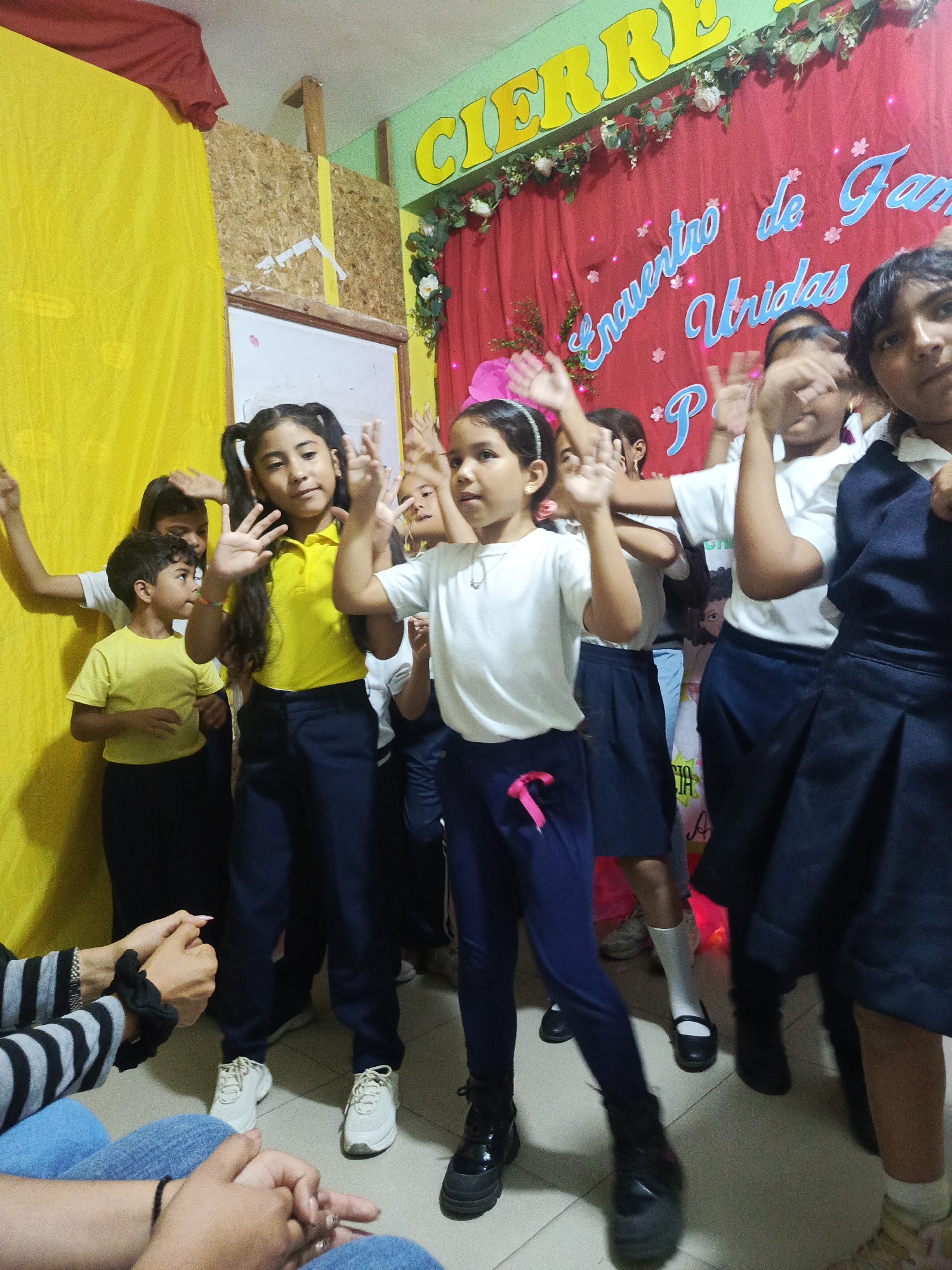 A group of elementary-aged children are gathered indoors, possibly in a classroom or community center. They are positioned in front of a red backdrop decorated with text that reads “Encuentro de Familias Unidas” (Meeting of United Families). The environment includes a yellow wall covering and floral decorations. The children, mostly wearing school uniforms of white shirts and navy bottoms, are actively raising their hands, seemingly participating in an activity or performance. In the foreground, the lower portion of seated adults wearing striped shirts and blue jeans is visible. The city or location is not evident based on the image alone, despite being in Juan Griego, Venezuela.