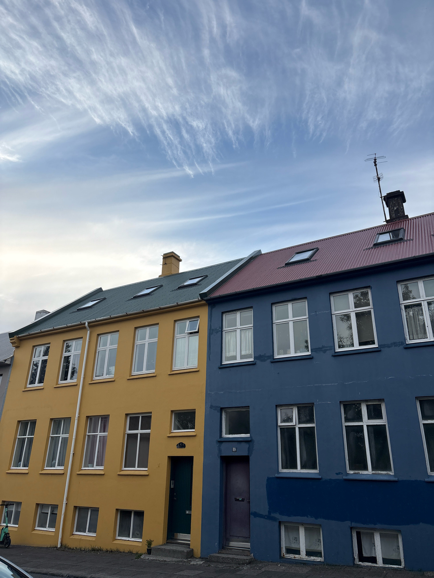 The image shows a side-by-side view of two residential buildings in Reykjavik, Iceland. The building on the left is painted yellow and has a green sloped roof with several skylights. The building on the right is painted blue and has a red sloped roof with skylights and a chimney with an antenna. Both buildings have multiple stories with numerous white-framed windows. The sky above is blue with thin, wispy white clouds. The lower part of the image shows a street with a sidewalk, a green electric scooter parked on the sidewalk, and the hood of a car in the foreground. The buildings appear to be attached and are situated in an urban environment.