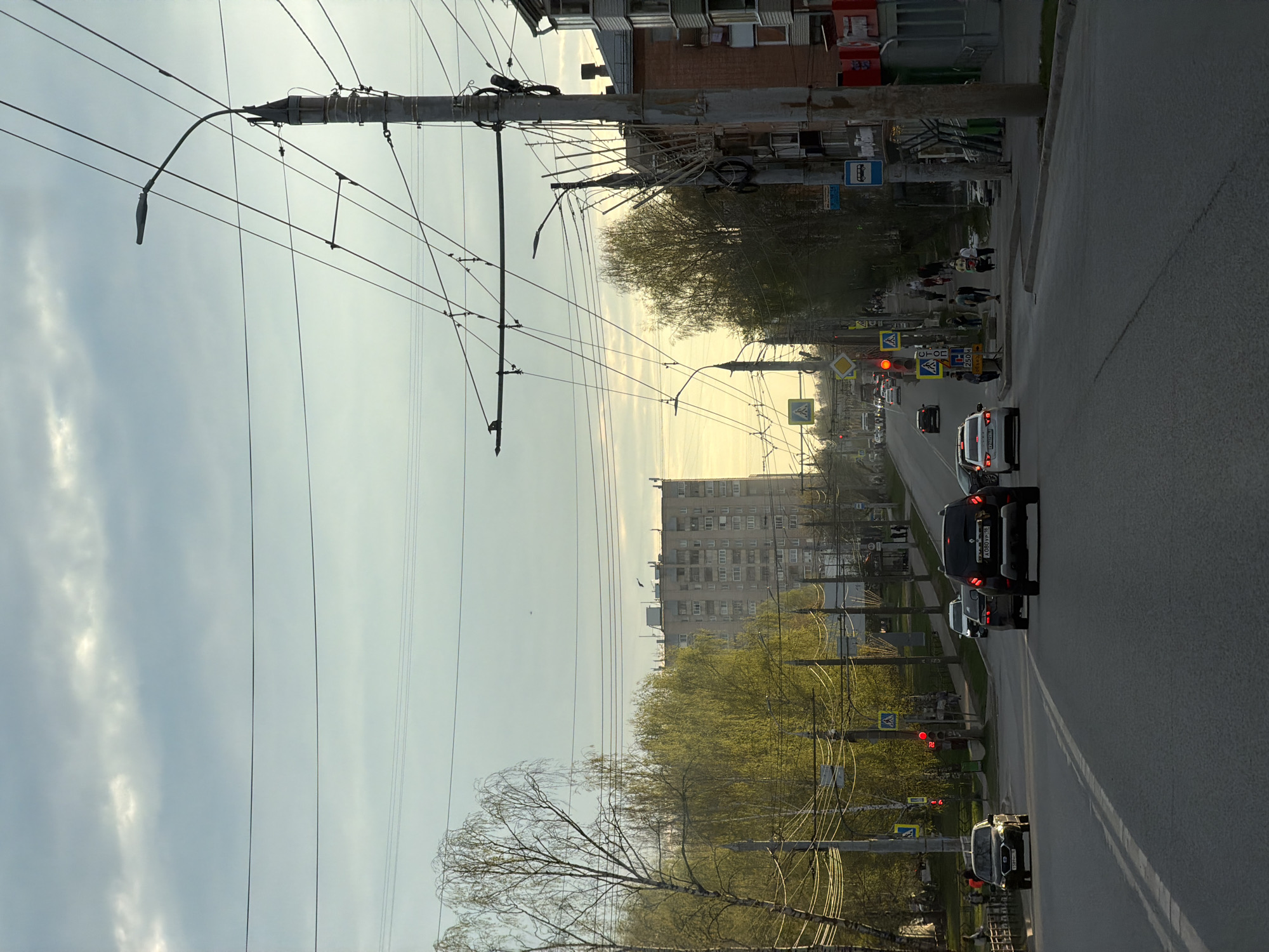 The image shows a street in Izhevsk, Russia. The street is lined with apartment buildings and trees. A row of cars is travelling along the road. Pedestrians walk alongside the street on the pavement. Traffic lights are installed along the street. Overhead power lines and trolleybus wires cross above the road. The sky is overcast.