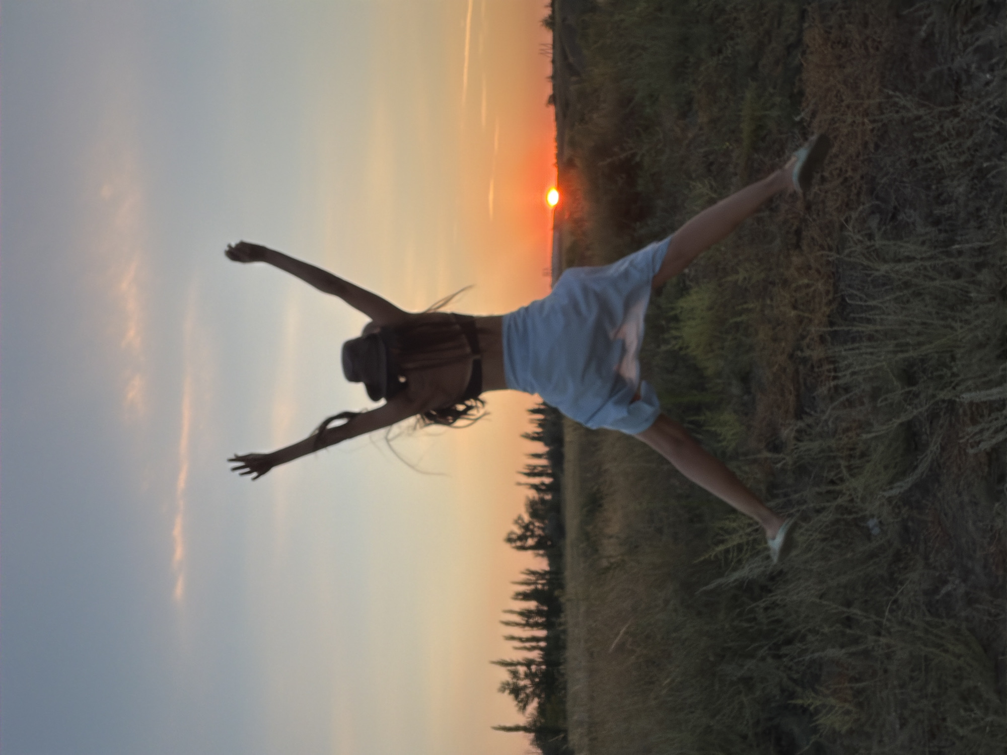 The image shows a person in silhouette jumping in a field at sunset. The person is wearing a hat and a light-colored skirt over shorts. Their arms are raised above their head. The sun is setting on the horizon, casting a warm glow over the scene. The field is covered in dry grass, and a line of trees is visible in the background. The sky is a gradient of blue and orange, with some wispy clouds. The location is likely in or near Kamyshin, Russia, given the landscape and the type of vegetation.