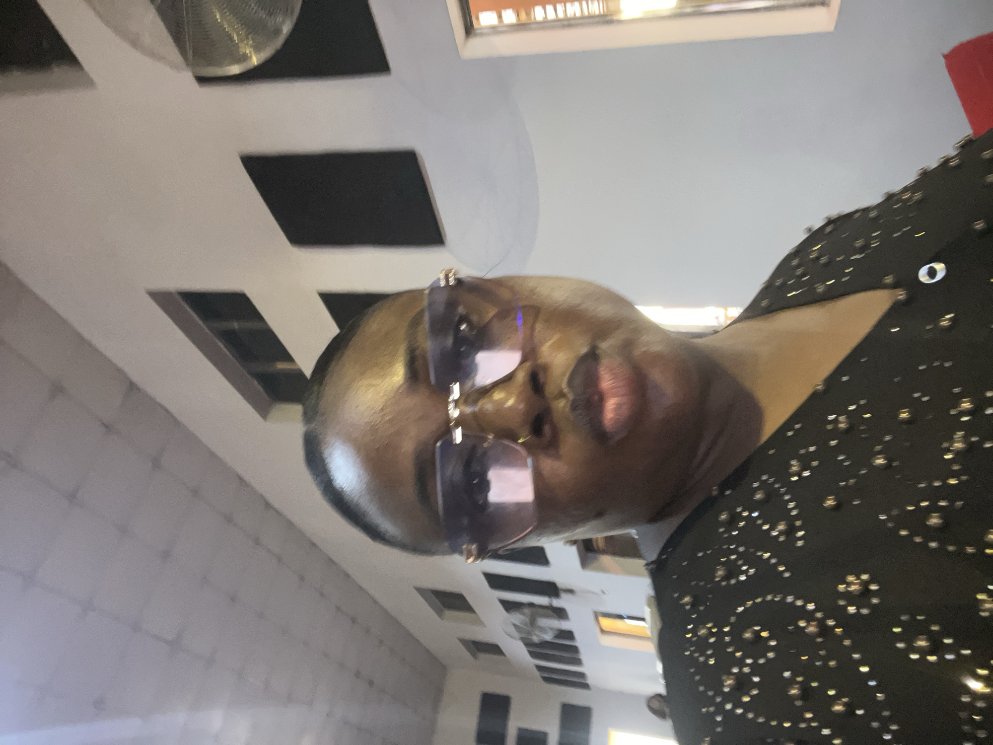 A person is pictured in a selfie. They are wearing sunglasses and a black, possibly beaded or studded top. The location is in a building with distinctive interior design elements including recessed rectangular features in the ceiling. Based on context clues available, the location may be in Abuja, Nigeria.