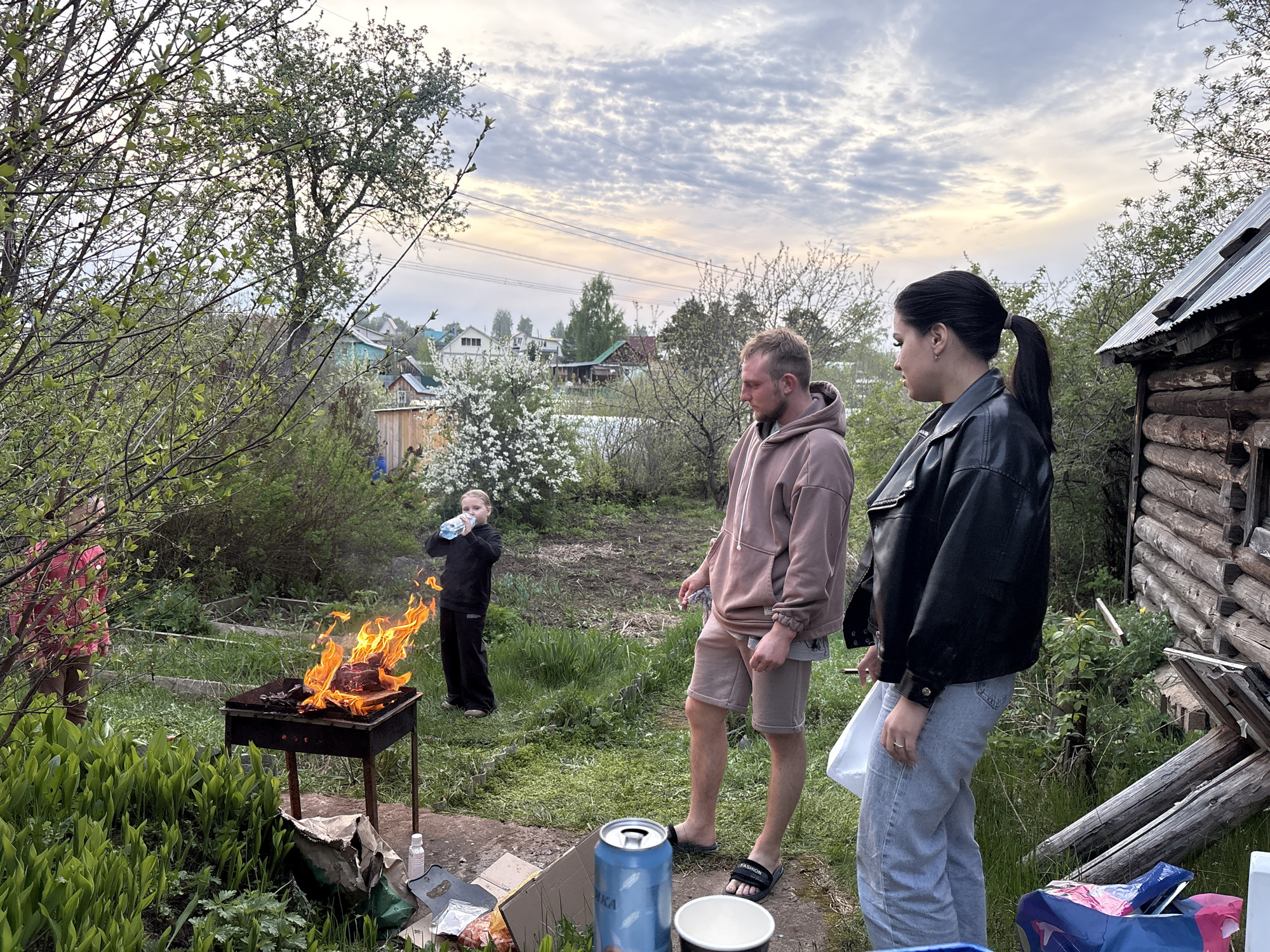The image, likely taken in Chaikovsky, Russia, shows an outdoor scene with three people near a barbecue. A fire burns in the grill, attended by a child in black. A man in a brown hooded sweatshirt and shorts stands nearby. A woman wearing a black jacket and jeans stands next to him. There is a can, a bowl, and a bag on the ground. In the background, there is a house and trees. The sky is cloudy.