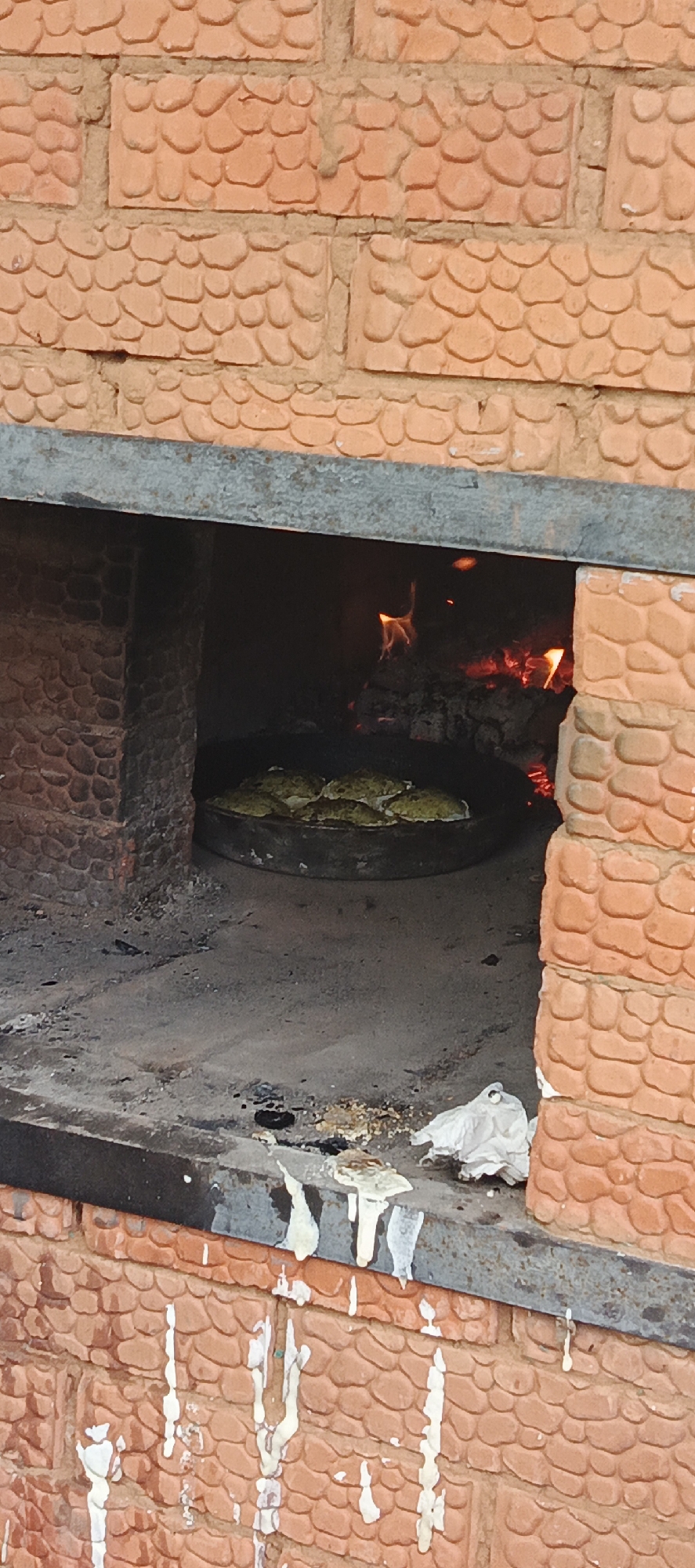 The image shows a close-up view of a rustic, brick oven used for cooking. The oven is constructed with textured, orange bricks that have a pebble-like pattern. Inside the oven, a fire is burning, with visible flames and glowing embers, indicating it's actively in use. In the foreground, a dark, round metal pan is placed on the oven's floor, containing several rounds of what appear to be baked goods, possibly bread or pastries, with a greenish hue. The floor of the oven and the hearth in front of it are dusty and show signs of ash and debris from previous fires. There are also some white paper-like materials, possibly used for cleaning or packing, resting on the hearth. The overall impression is one of a traditional, perhaps rural, cooking setup in Старые Быги, Russia. The lighting suggests it could be daytime, but the interior of the oven is dominated by the warm glow of the fire. There is no visible text in the image.