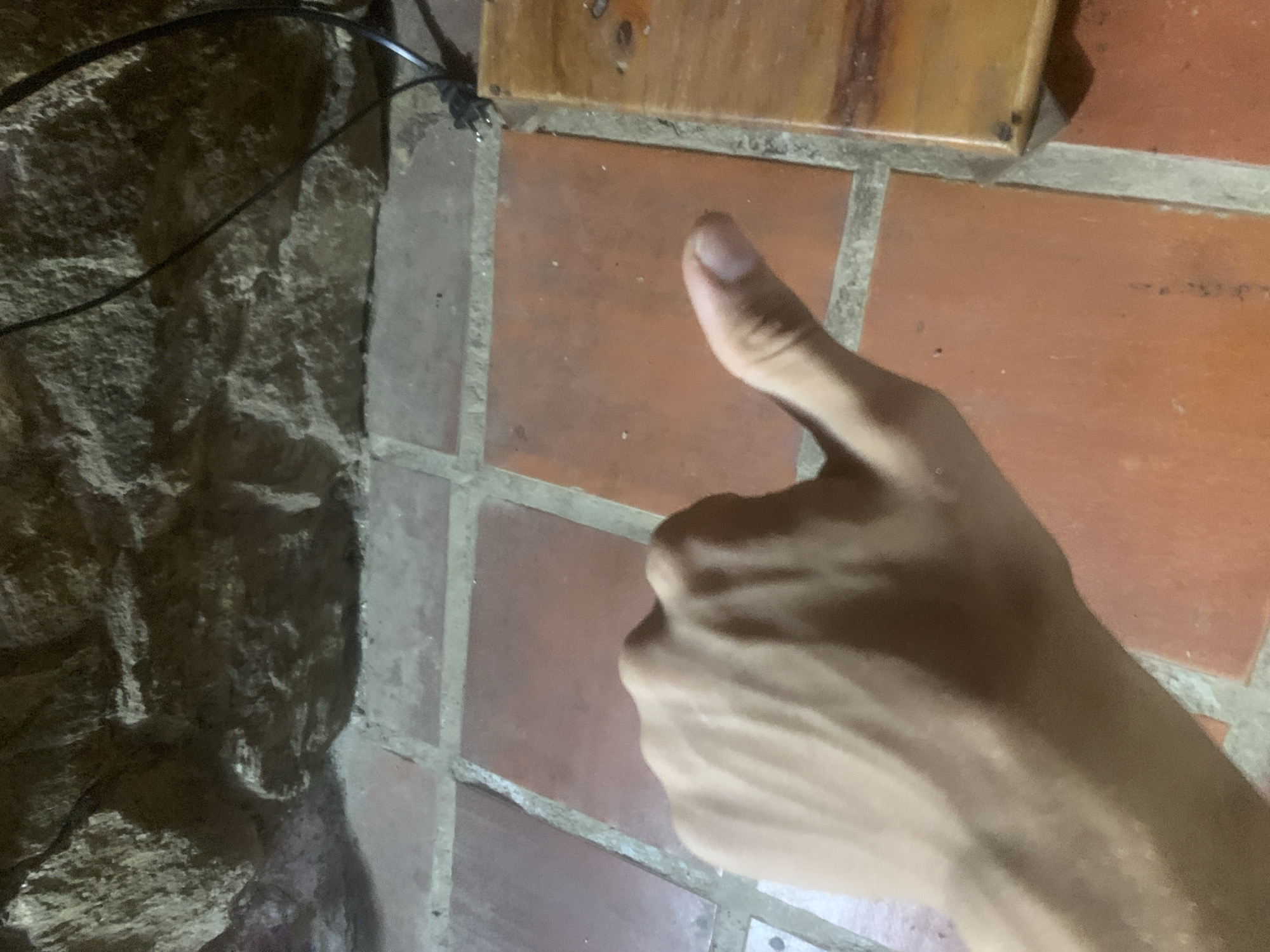 A person's left hand with prominent veins is shown extended forward, with the thumb pointing upwards in a "thumbs-up" gesture. The hand is positioned in the foreground against a tiled floor made of reddish-orange square tiles with visible grout lines. Behind the hand, a rough, textured stone wall forms the background. To the right of the hand, a portion of a wooden object is visible, possibly a piece of furniture, with some dark smudges on its surface. A black electrical cord is also present, originating from the upper right and trailing down towards the lower right corner of the image. The overall scene appears to be an indoor or semi-outdoor setting with a rustic aesthetic. The location of Pedro González, Venezuela, is not discernible from the visual information provided in the image.