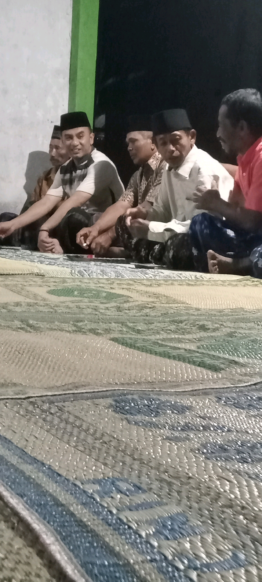 In Kedungmulyo, Indonesia, five men are seated on a woven mat, likely for a gathering or meeting. The men are all wearing traditional attire, including peci caps. One man in the center is holding a lit cigarette, and another man to his left is also holding a cigarette. The environment appears to be outdoors, possibly a courtyard, with a wall visible in the background. The lighting suggests it is evening or night. The woven mat on which they are seated has a patterned design.